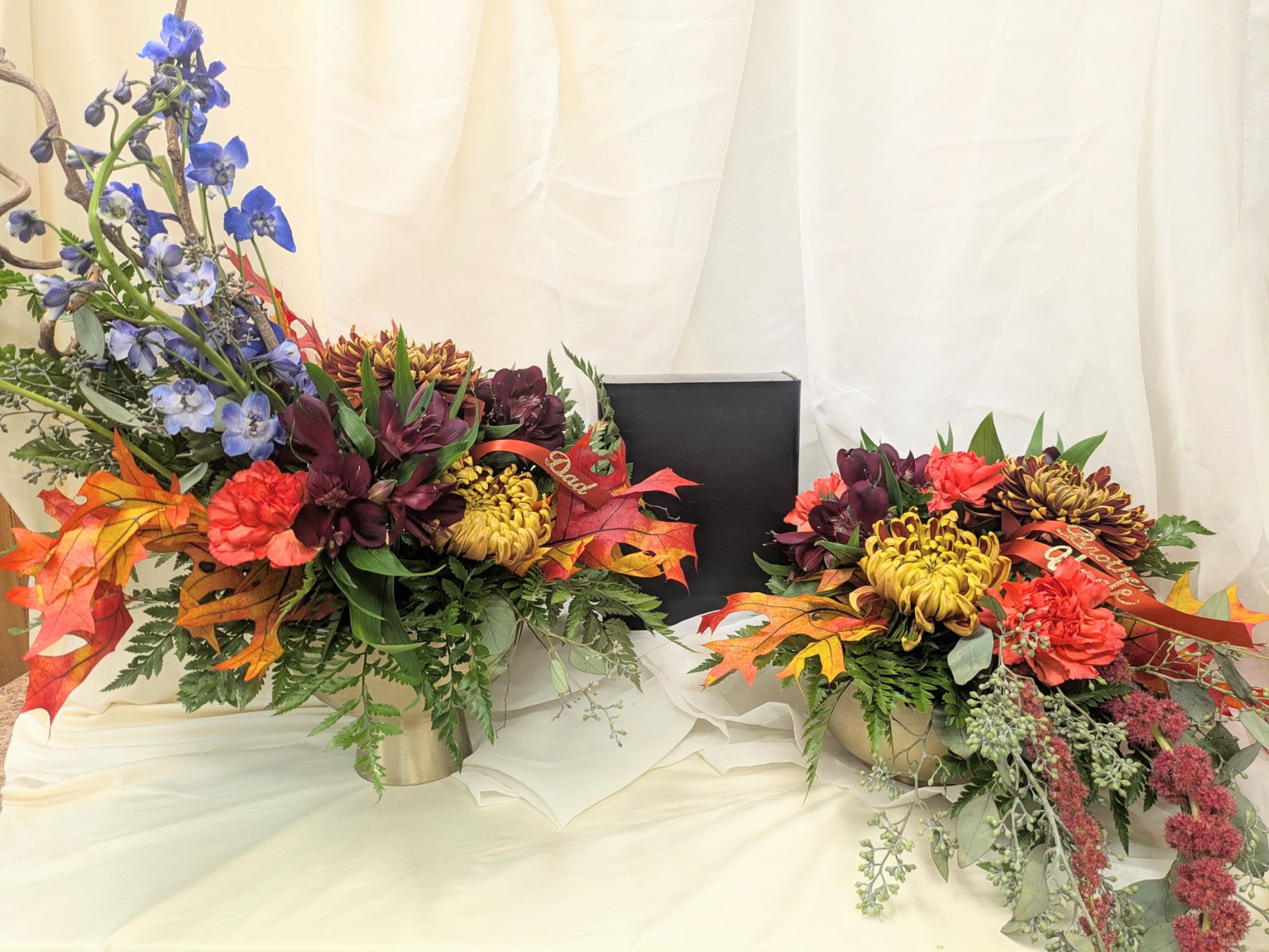 Eternal Harvest Urn Adornment  - A stunning tribute inspired by the beauty of fall. The Autumn Memories Urn Adornment features rich tones of burgundy, gold, rust, and deep blue — symbolizing warmth, strength, and enduring love. Accented with seasonal leaves and lush greenery, this arrangement beautifully frames the urn and offers a heartfelt expression of remembrance and peace.  Disclaimer: This design is intended as an urn adornment, created specifically to accompany and enhance a loved one’s urn during a memorial or celebration of life service. It is not meant to be sent as a sympathy arrangement or delivered directly to family members. If you wish to send flowers in remembrance or as a gesture of comfort to the family, please select from our sympathy or memorial floral arrangements instead.  All flowers, flower colors, and containers are subject to change due to availability. 