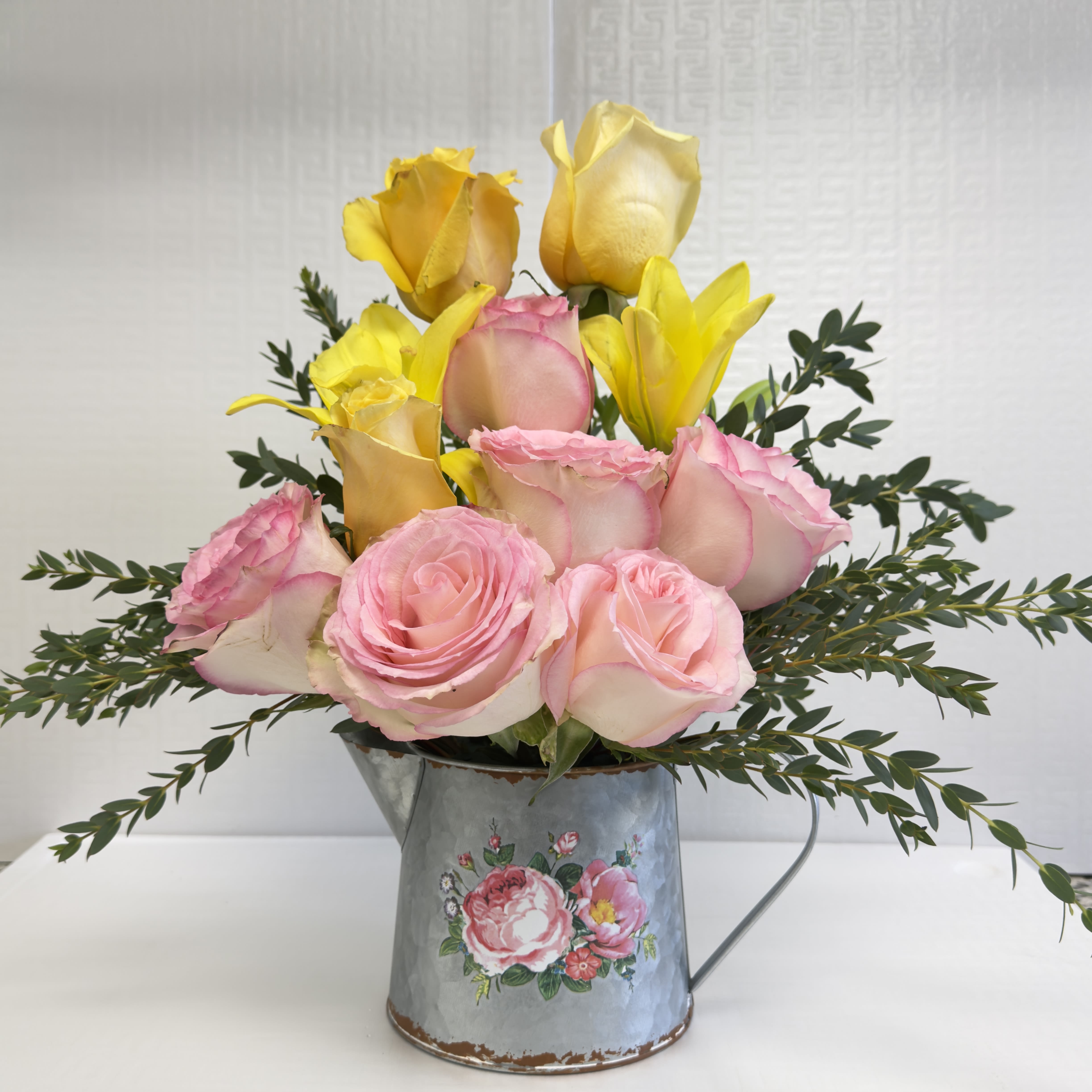 Sunshine Bliss -  A cheerful mix of pink and yellow roses with yellow lilies arranged in a charming vintage-style watering can. This bright and joyful design brings a touch of sunshine and sweetness to any space — perfect for birthdays, thank-yous, or just because.