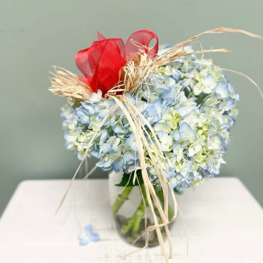 Turning Point - Turning Point” pays tribute to the life and legacy of Charlie Kirk — a figure who challenged young people to stand courageously for their beliefs and reimagine the direction of their futures. This arrangement brings together serene blue hydrangeas paired with a vibrant red ribbon and rustic raffia accents.  It’s more than a floral piece: it embodies resolve, patriotism, conviction, and the moment when one decides that change is essential. Let Turning Point be a symbol of remembrance, inspiration, and the hope that bold ideas and faith in one’s values can light the path forward.