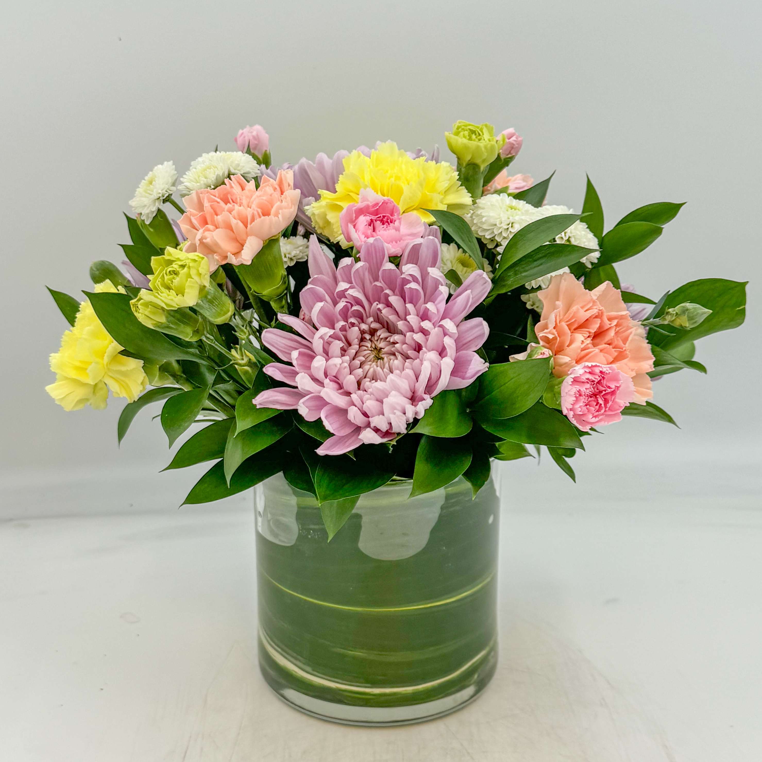 Nonna Primavera - My Nonna absolutely hated receiving flowers that were not mums or carnations because they live the longest. Therefore, Nonna Primavera is the perfect long lasting spring arrangement, designed with Lavender Chrysanthemums, White Button Mums, and Yellow and Peach Carnations, Pink and Green Mini Carnations in a bed of Israeli Ruscus and an Aspidistra Leaf liner. Measurements: The Standard and Deluxe Nonna is designed in a 5x5 inch glass cylinder vase and the Premium Nonna is designed in a 6x6 inch glass cylinder vase.