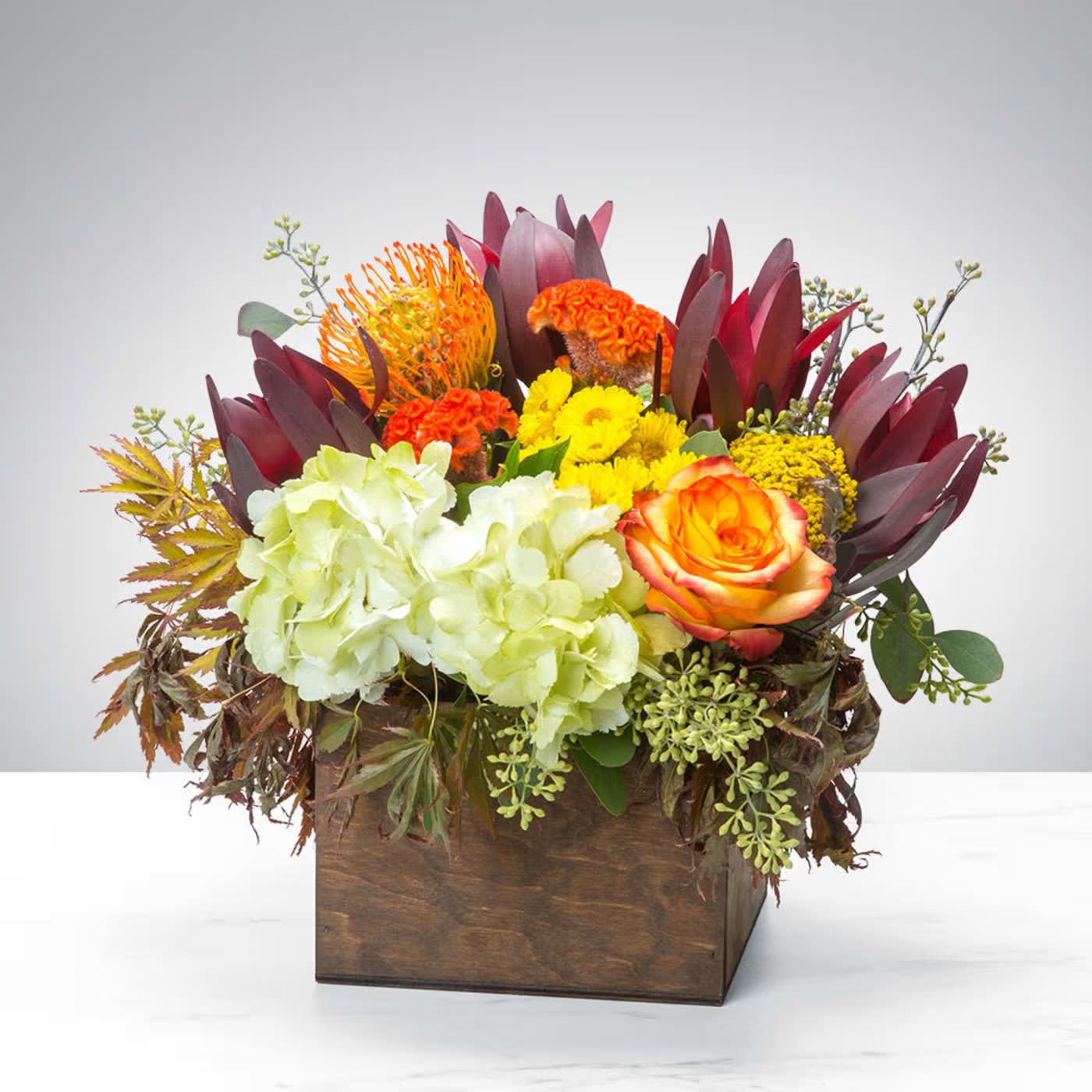 Autumn Celebration ( BB ) - Introducing our enchanting Flower Arrangement in a wooden box, a captivating combination of green hydrangeas, orange roses, yellow buttons, seeded eucalyptus, and more. This arrangement is carefully curated to create a stunning display of contrasting colors and textures, housed in a rustic and charming wooden box.  