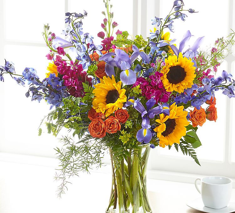 Summer Skies - Show your loved ones how much you care with this bright and beautiful arrangement of summer flowers including sunflowers, stock, iris, roses and or spray roses and delphinium. This bouquet will include bright vibrant summer colors like fuchsia , blue, yellow -gold, orange, purple, and bright green.  What a way to brighten someone's day or you just to say hi,  this arrangement is sure to be great for any occasion. 