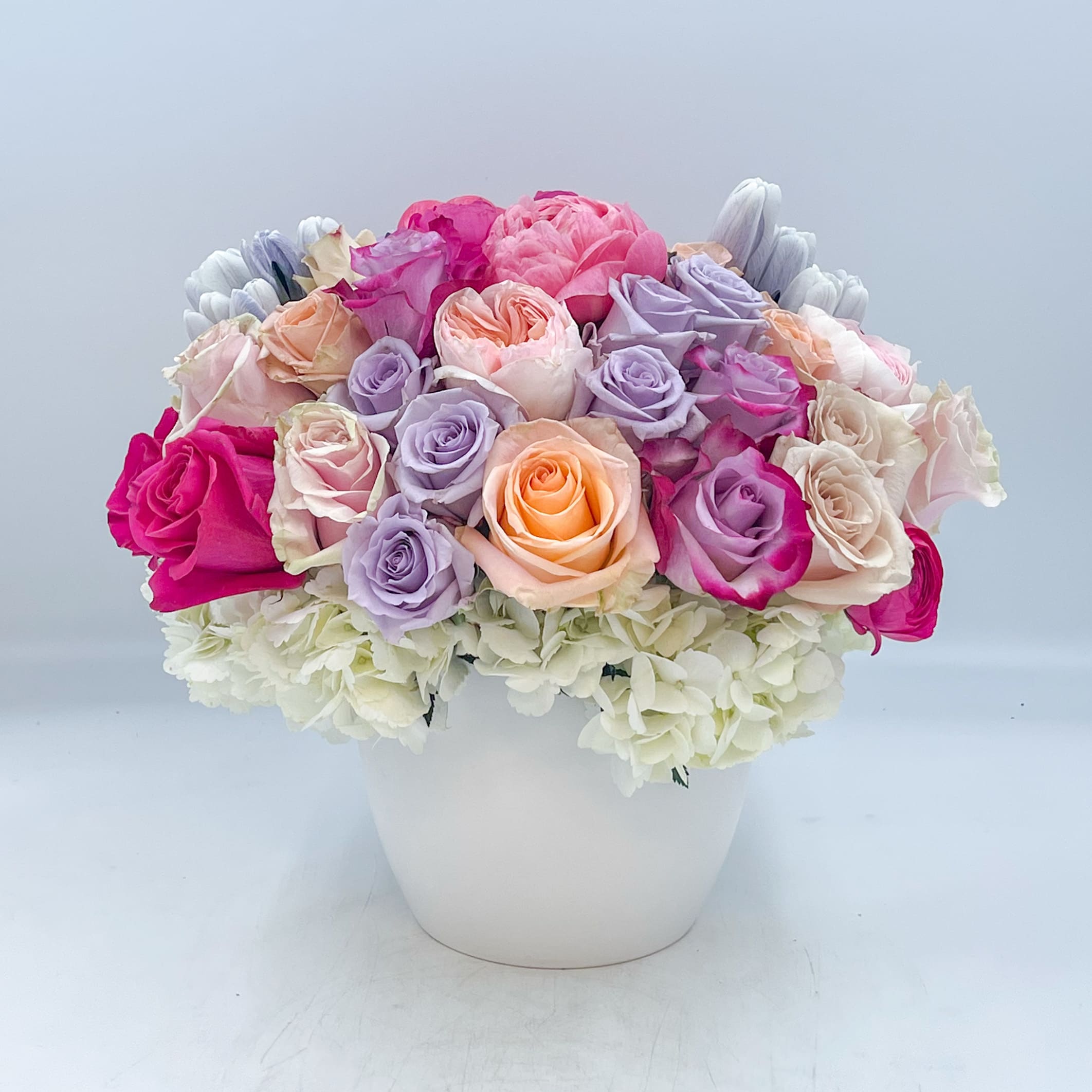 Dixie - Dixie is one of our most upscale and premium designs. Dixie includes White Hydrangea, Coral Charm Peonies, Julie Garden Roses, Lavender Roses, Peach Roses, Blush Roses, Hot Pink Roses, Quicksand Roses, Blue Maggie Tulips, Blush and Hot Pink Italian Clooni Ranunculus designed in a 6 inch White Ceramic Container. 