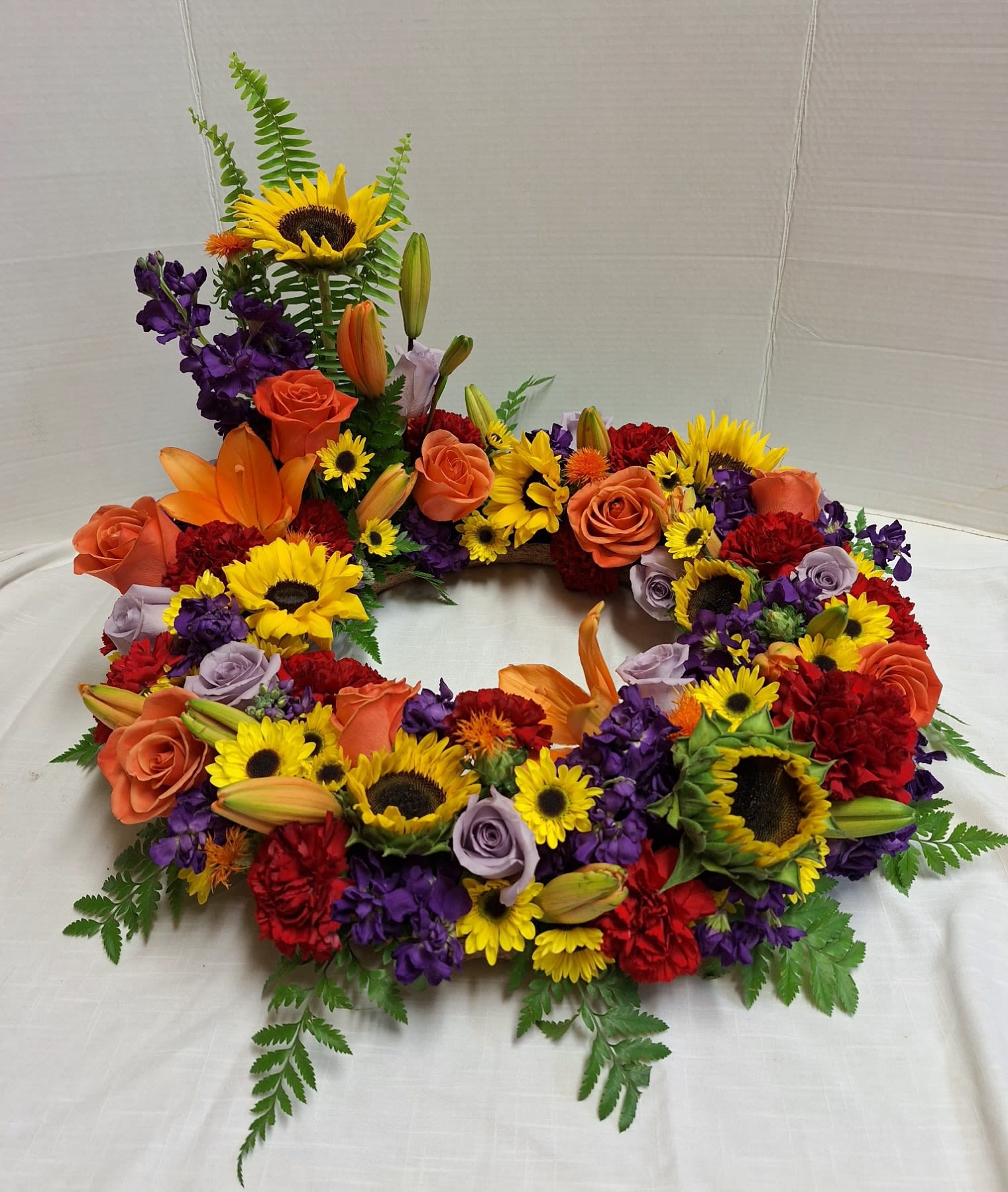 Autumn Burst Urn Wreath - Brilliant burst of color. Orange and Lavender Roses. Orange Lily’s, Red Carnations and assorted size Sunflowers. Along with Purple Snap Dragons.