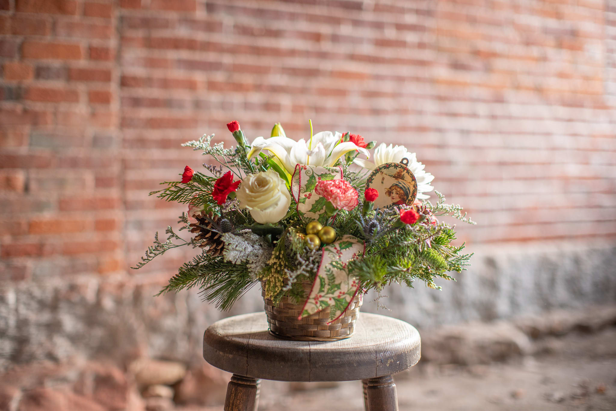 Vintage Wishes Basket - Adorable vintage take on the whimsical winter tales of past.  Winter foliage encompasses a wooden pick with classic graphics of a snowmen, Santa, and other seasonal scenes. Beautiful blooms and matching ribbons come to life like the magic in his hat! Blooms include standard and mini carnations, sweet smelling lilies and a rose. Lilies may arrive in bud stage but will open beautifully in a trumpet shape graduating to full openness of a star.