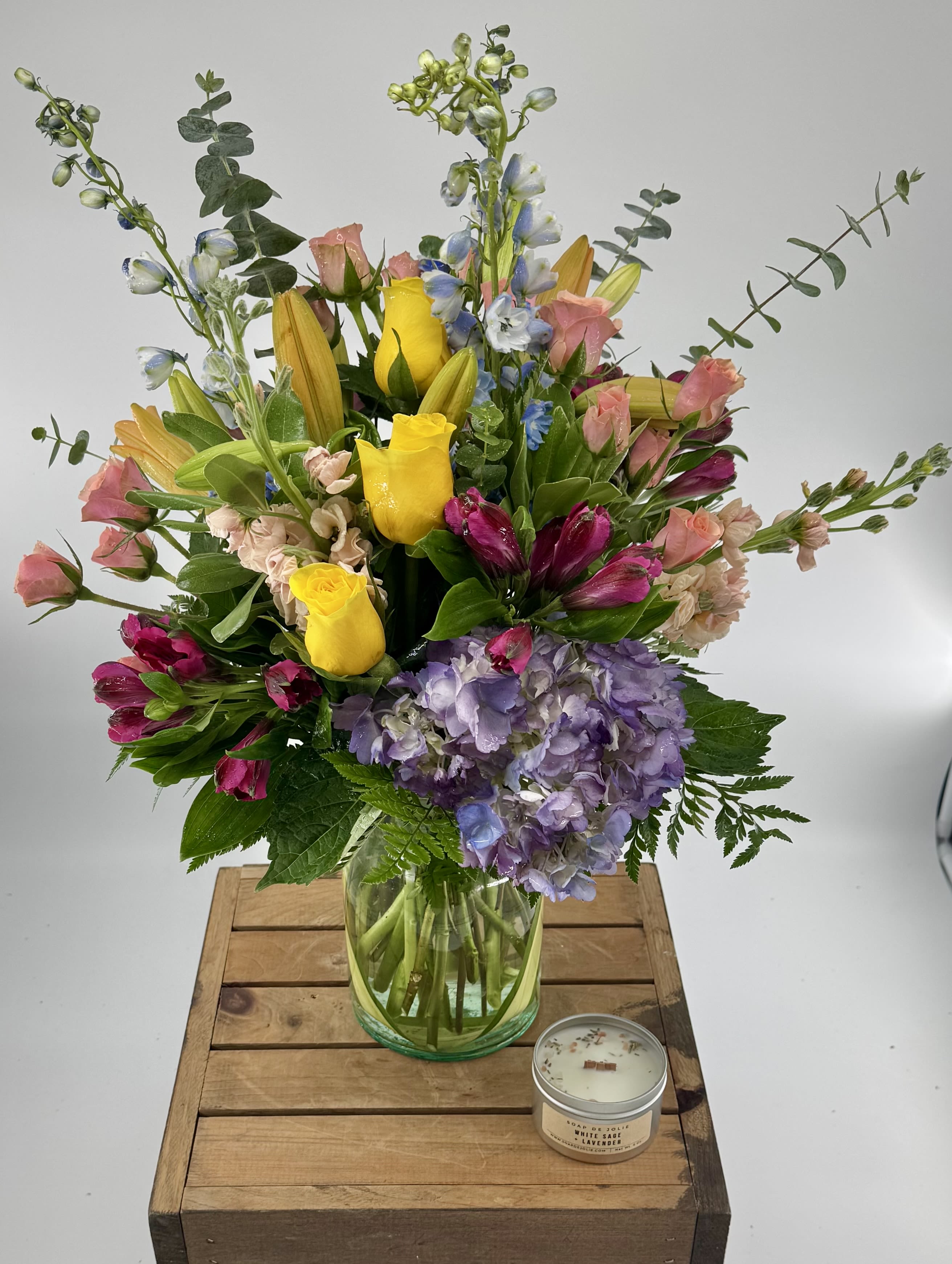 Happy Days  (BB) - Elevate your space with our modern floral arrangement, thoughtfully crafted in a clear glass leaf-lined vase. This artistic ensemble features a vibrant mix of orange lilies, delphinium, stock, spray roses, purple hydrangea, yellow roses, and assorted greenery. Captivating in its design, this arrangement brings a burst of color and elegance to any room, making it a perfect statement piece for any occasion. 