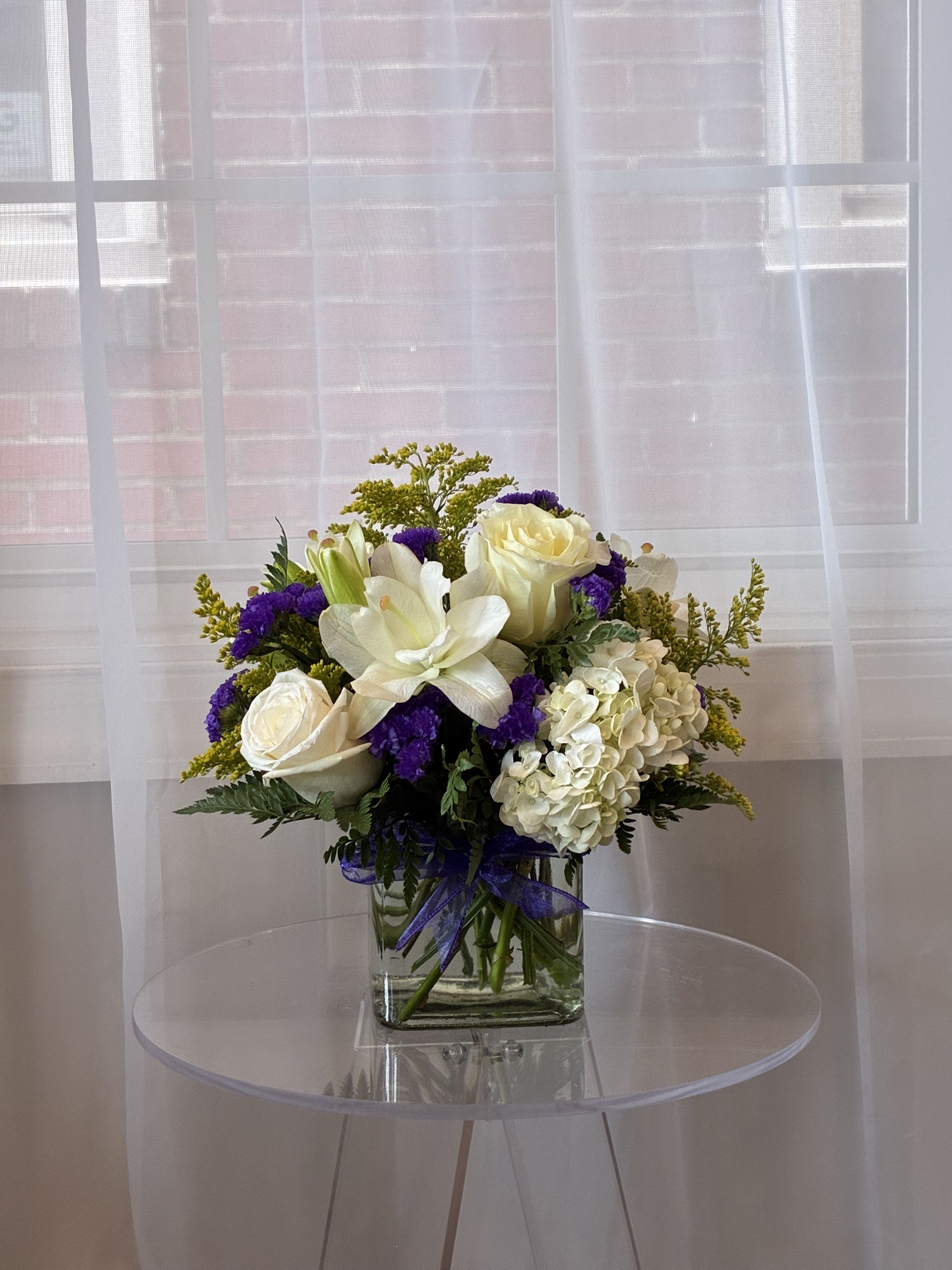 Simple Song - Bring a calming elegance into any space with our Simple Song arrangement, a hand-designed floral display featuring a soft, refreshing color palette of purples, whites, and yellows. This calm bouquet includes white hydrangea for a voluminous, serene base; white Lilies for their luxury, timeless charm; white roses to symbolize grace; Yellow solidago to provide height &amp; texture; and purple statice for a delicate, fragrant texture. Seasonal greenery adds fullness and contrast to complete the look.  Artfully arranged in a glass cugbe, this design is perfect for sympathy expressions, spring celebrations, or simply to brighten someone’s day with a sophisticated touch of nature.