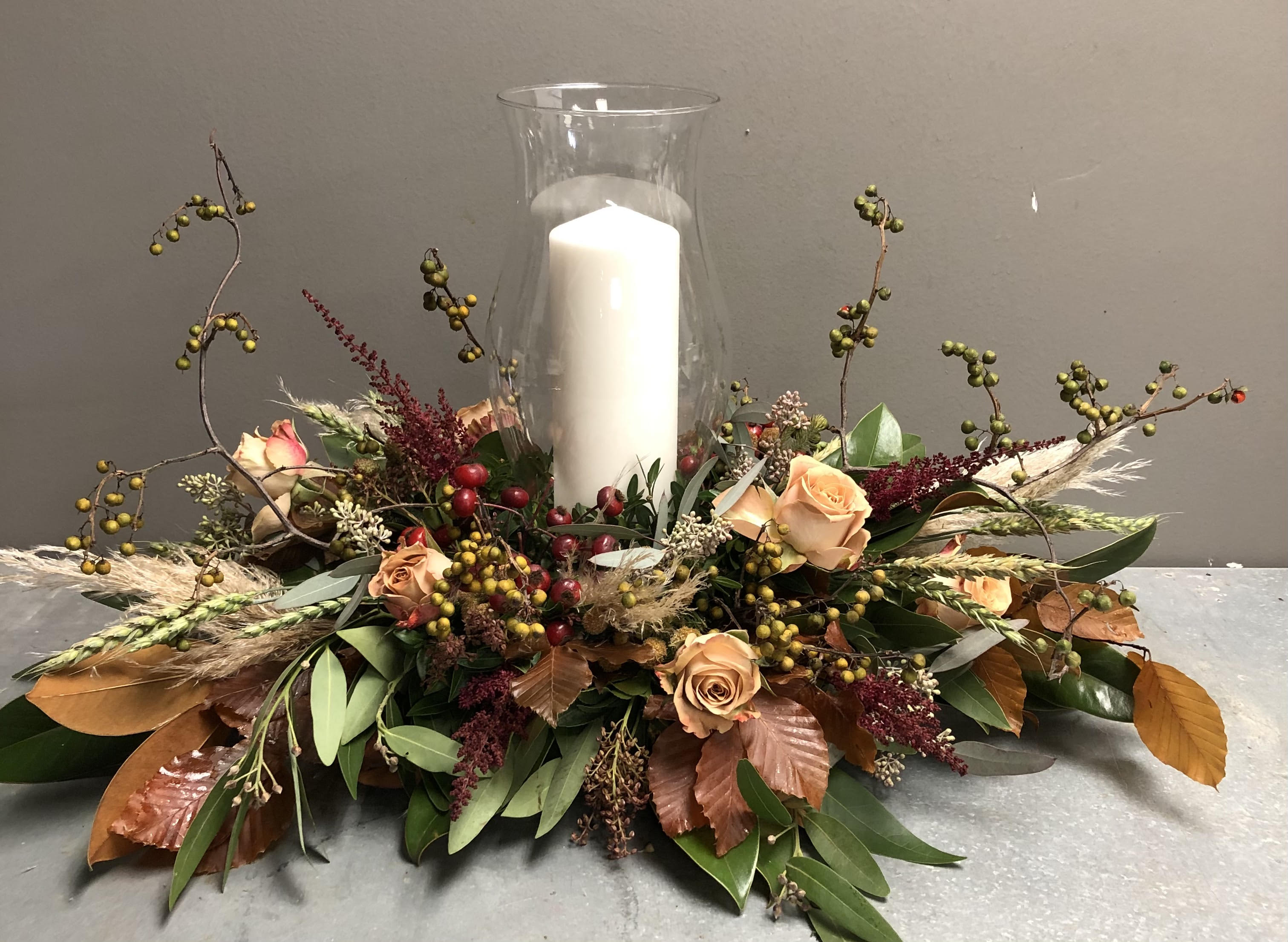 HARVEST MOON - Gather family and friends around your most beautiful table with this abundant Thanksgiving Centerpiece. Honey-hued roses are shown with richly textured elements, centered with a glass incased pillar candle, and seasonal berries. 