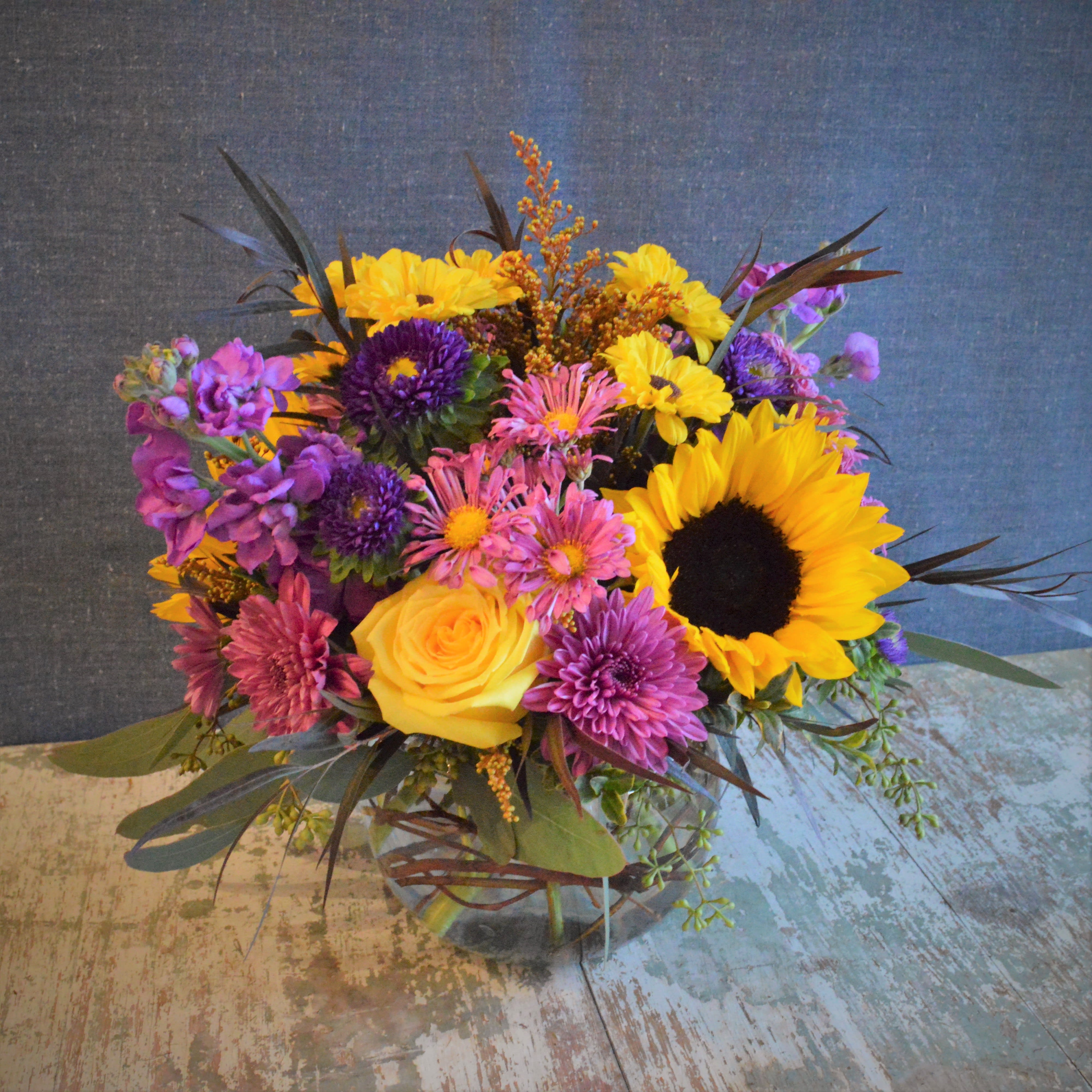Golden  - Seasonal flowers in complimentary colors of purple and yellow with dark foliage accents. This arrangement is designed in a curly willow lined bubble bowl. Approximately 12&quot; tall by 10&quot; wide. 