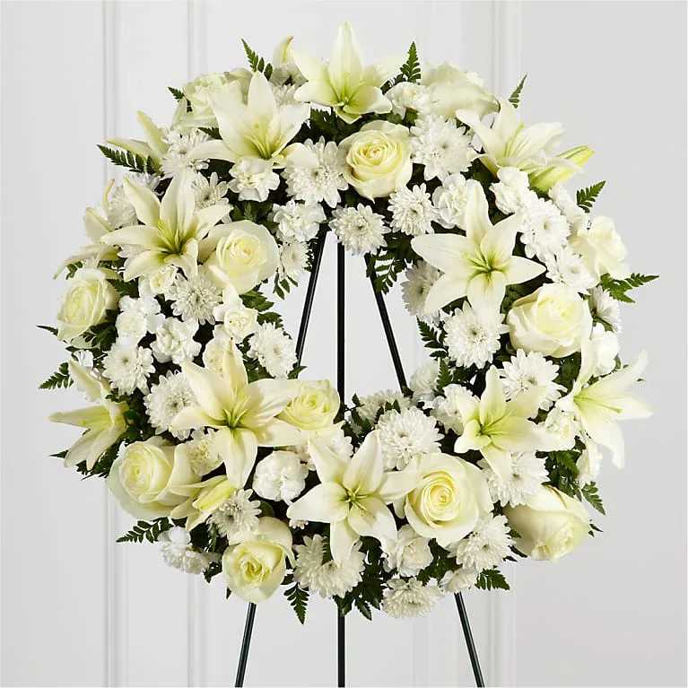 FTD TREASURED TRIBUTE WREATH - TREASURED TRIBUTE WREATH  The Treasured Tribute Wreath offers peaceful wishes of heartfelt sympathy with each delicate bloom. Bright white roses, Asiatic lilies, mini carnations and cushion poms are beautifully arranged to form an elegant wreath accented wth lush and vibrant greens. Displayed on a wire easel, this gorgeous tribute is a wonderful symbol of eternal life and sweet serenity.