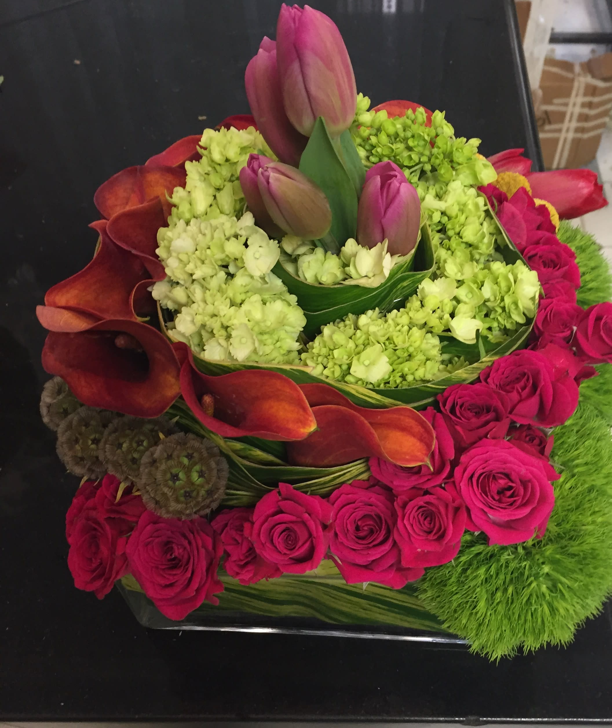 Crimson Garden Luxe - A striking modern composition featuring rich crimson roses, vibrant green hydrangea, and velvety orange calla lilies layered in sculptural harmony. Accented with fresh tulip blooms and textured green trick dianthus, this square glass design is wrapped with folded ti leaves for a polished, contemporary finish.  Perfect for luxury celebrations, corporate gifting, or a statement centerpiece — this design blends high-end florals with architectural artistry, creating an unforgettable visual impact from every angle.