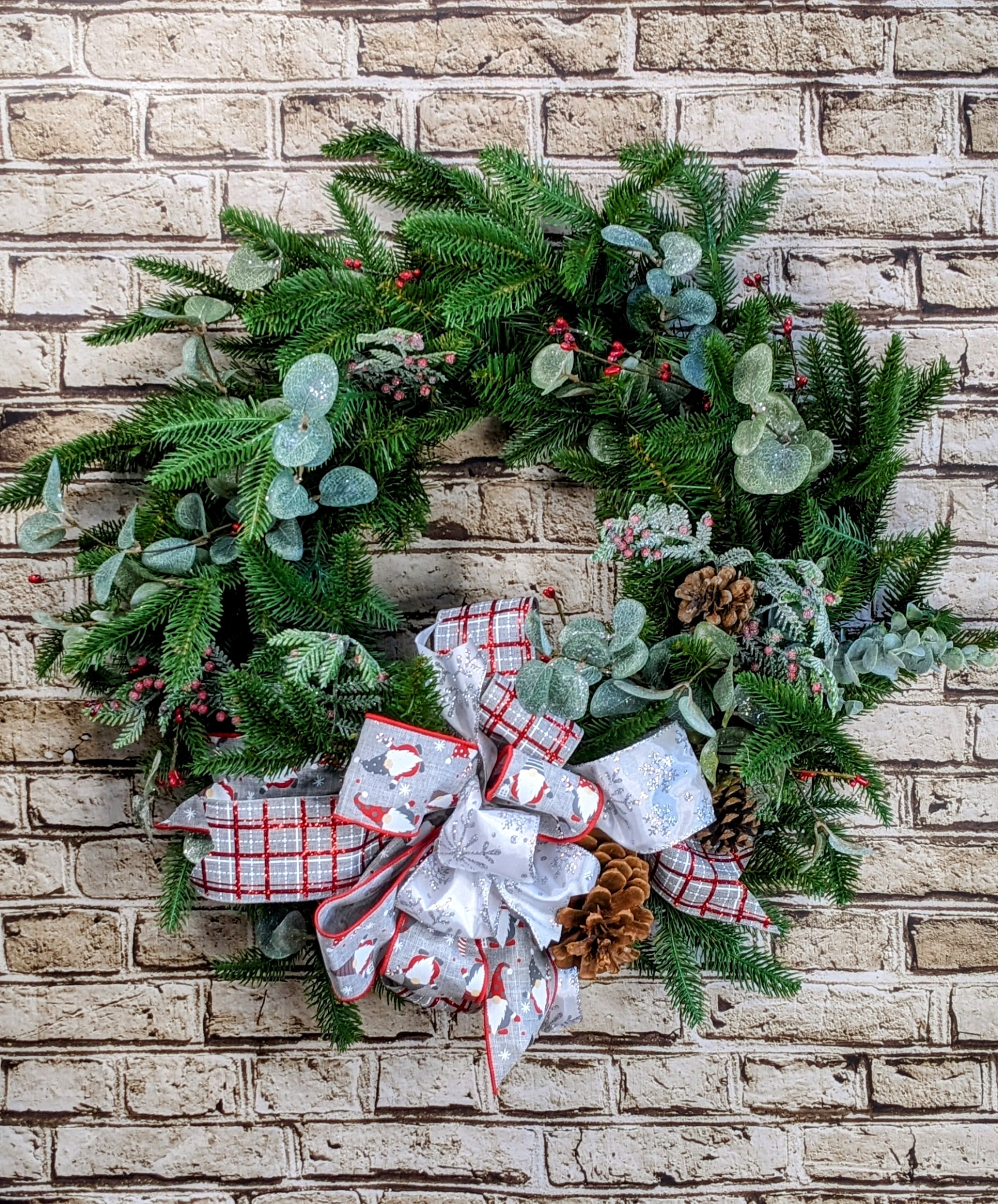 Seasonal Holiday Wreath - 24&quot; Artificial - A classic 24-inch artificial holiday wreath, exuding timeless charm with its lush greenery and subtle eucalyptus accents. Red and silver embellishments add festive color, while matching ribbon weaves through the design for a coordinated, elegant look. Rustic pinecones are thoughtfully placed throughout, enhancing the natural appeal. This wreath is perfect for creating a welcoming, seasonal atmosphere with a sophisticated blend of traditional and modern elements.  Approximate Dimensions: 24&quot; Diameter