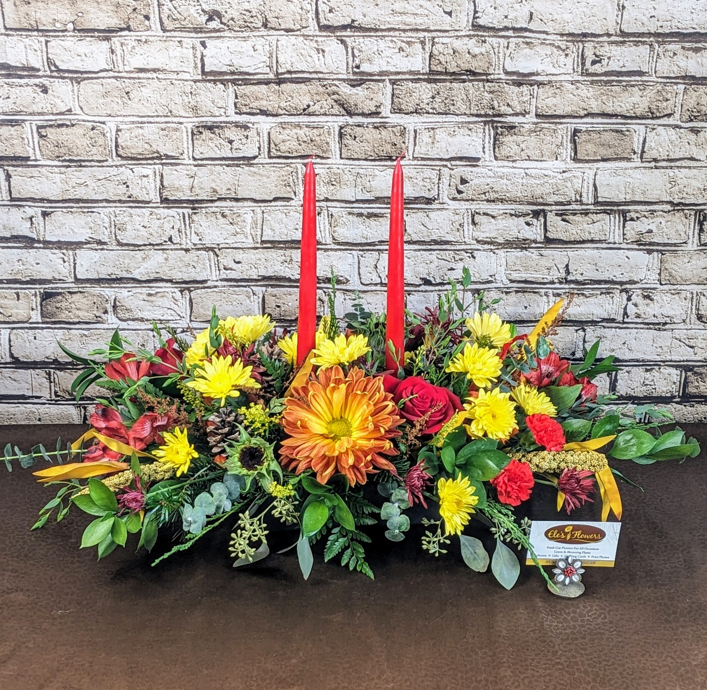 Vibrant Thanksgiving Centerpiece - A vibrant Thanksgiving centerpiece with a rich autumnal palette features a harmonious blend of bright yellows, fiery oranges, and deep reds. The arrangement incorporates natural accents such as pinecones, dried leaves, and sprigs of fragrant greenery, adding texture and an organic feel. Two elegant red candles stand tall at the center, their warm glow enhancing the festive atmosphere.   Centerpiece will arrive similar as shown, some blooms may vary.  The designs we create are custom and the blooms and containers will vary. We use the freshest product available to create the perfect floral combination for the recipient. If you have any special requests, please add those in the &quot;special instructions&quot; portion of your order to help us dial in your vision. Otherwise, our skilled designers, Sara &amp; Brett, will take it from here!  The design is a perfect for long gathering tables that seat 10 -12 people. Approximate Dimensions: 32&quot;L x 15&quot;H(including candles) 
