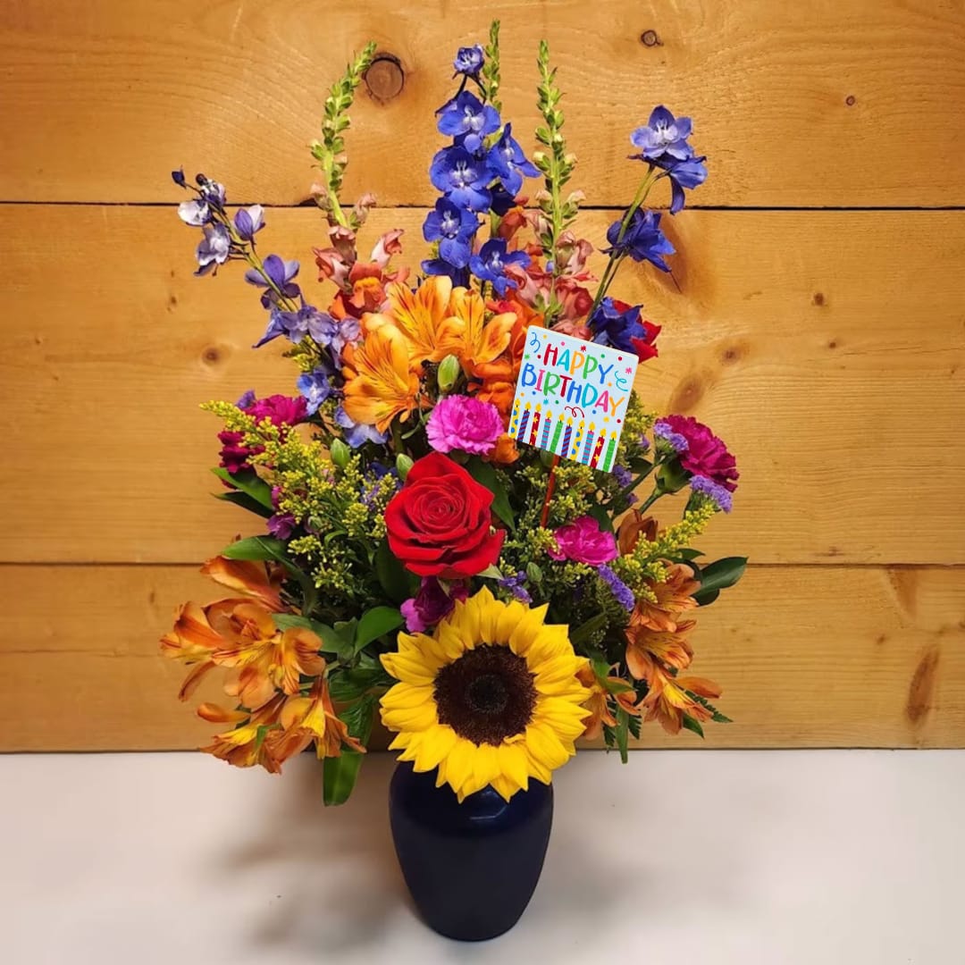 Birthday Radiance - Experience the warmth and joy of a perfect day with the &quot;Birthday Radiance&quot; floral arrangement, a dazzling bouquet that radiates cheer and elegance. Nestled in a striking blue vase, this arrangement showcases the sunflowers' golden glow, symbolizing happiness and positivity. Delphinium adds a touch of grace with its tall, enchanting spikes of blue, perfectly complementing the vase. Snapdragons bring vertical interest and a playful spirit with their vibrant colors and unique shape. Mini carnations and carnations offer a soft, lush texture in varied shades, while Alstroemeria adds a delicate, exotic flair. Solidago introduces a burst of golden brightness, and statice provides a subtle, airy texture that enhances the bouquet's overall charm. Together, these flowers create a harmonious and uplifting display, perfect for celebrating the special occasion with a touch of birthday brilliance.