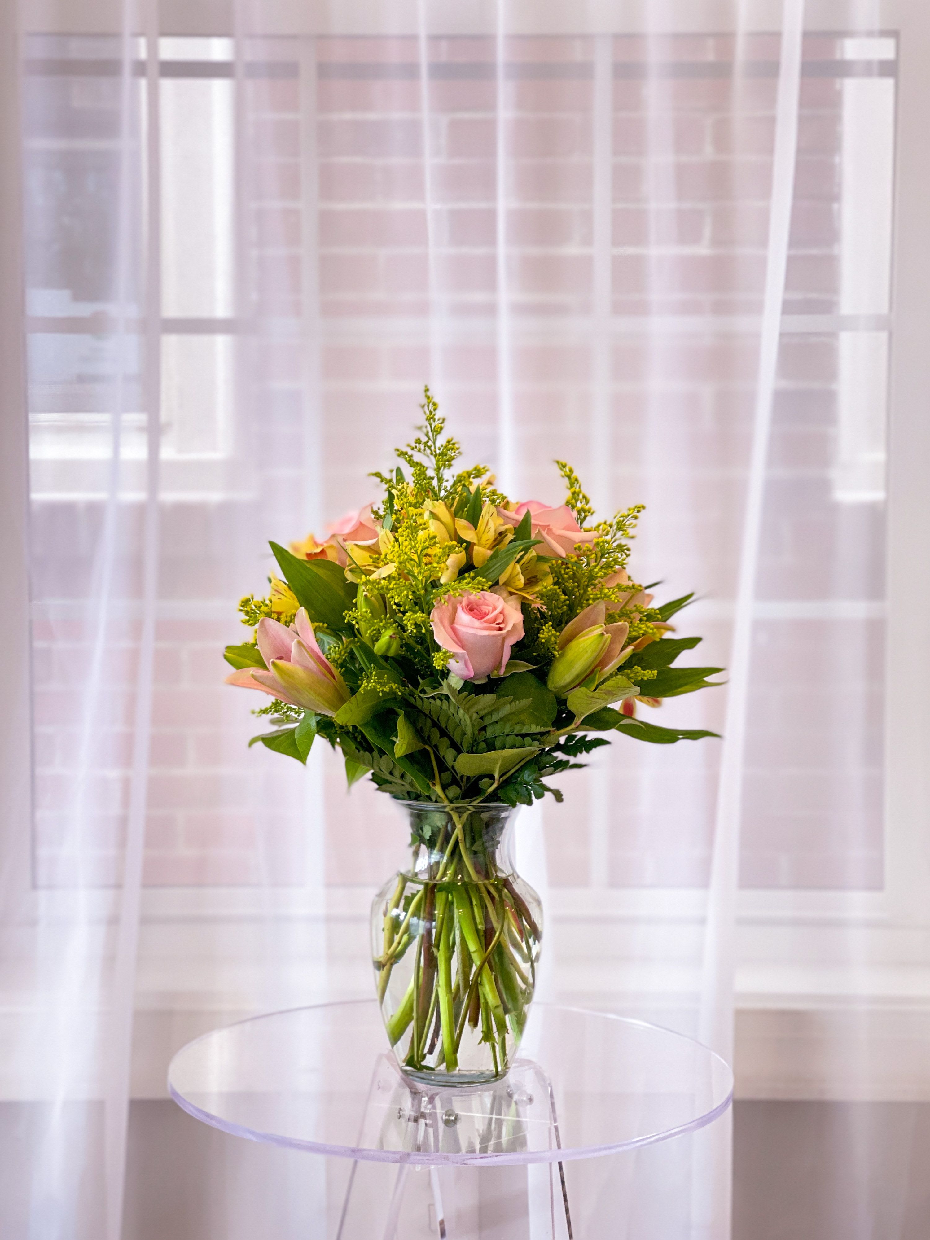 Sweet Bloom  - A soft, romantic bouquet that brightens any room. Pale pink roses nestle among fresh lily buds, airy golden solidago, and lush seasonal greenery. Arranged by hand in a clear glass vase, the clean silhouette lets the blooms take center stage while long, graceful stems add modern elegance. Perfect for birthdays, thank-yous, or “just because.”