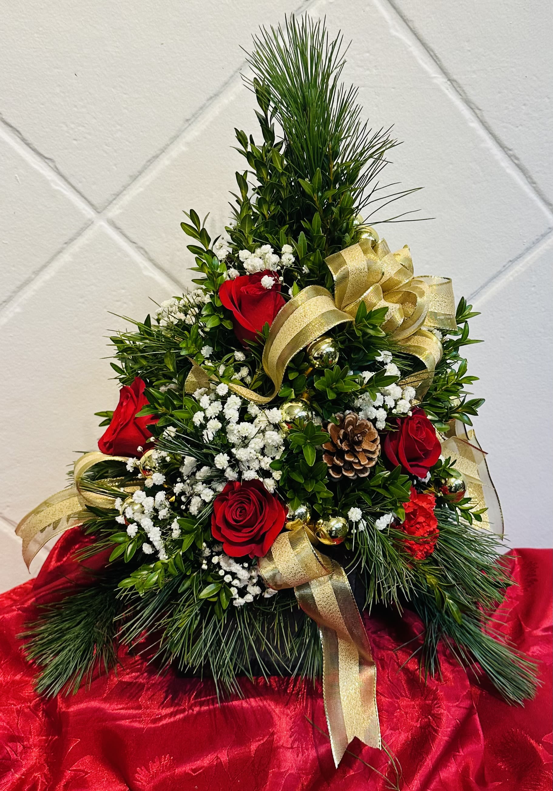 Crimson Joy Holiday Tree - The Crimson Joy Holiday Tree is a stunning centerpiece that promises to be the crown jewel of holiday decor. Crafted with care, it features lush pine and verdant boxwood shaped into a charming miniature tree, forming a verdant canvas for the vibrant hues that adorn it. Bold red roses and carnations are tucked among the greenery, their petals as rich and inviting as the season itself. Baby’s breath sprinkles the arrangement like delicate snowflakes, while shiny baubles peek out like hidden treasures. Golden ribbons weave through the branches, adding a touch of opulence and mimicking the cascading lines of tinsel. This arrangement not only delights the eyes but also fills the room with the heady, sweet scents of pine, creating an ambiance of warmth and festive elegance. It is a testament to the beauty and fragrance that embody the joy and essence of the holiday season.