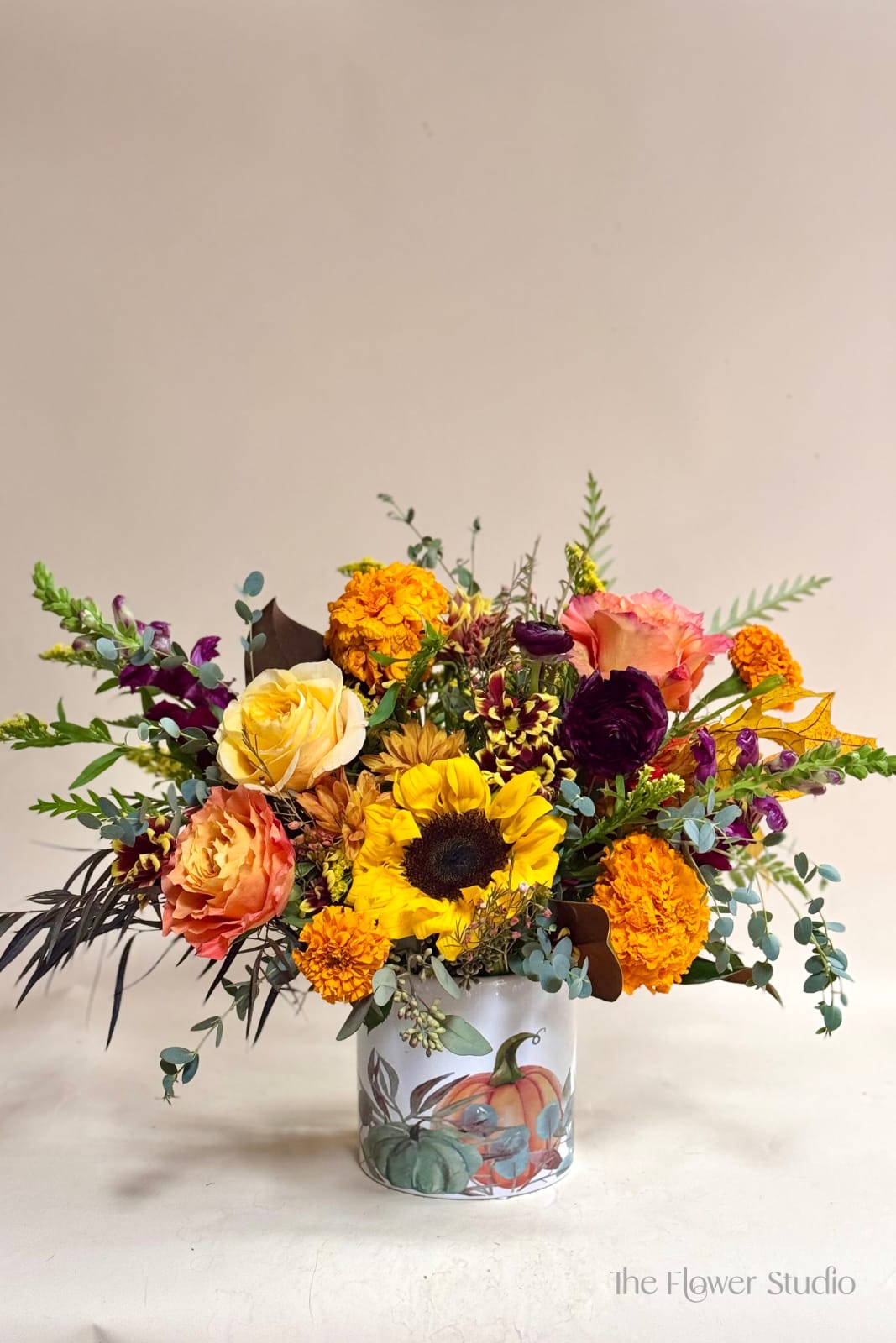 Fall in Love - A cheerful burst of fall color featuring bright sunflowers, peach and yellow roses, and rich burgundy blooms, all artfully arranged with lush seasonal greenery. Designed in a charming pumpkin vase, this arrangement perfectly captures the warmth and beauty of autumn. Ideal for a Thanksgiving centerpiece or to bring a cozy seasonal touch to any home. Approx: 20” W x 16” H Our floral artists hand-design each arrangement, so flower and container styles may vary based on local and seasonal availability. As a result, we are unable to guarantee specific florals, colors, or vessels, but we always ensure the same level of beauty and value in every piece.