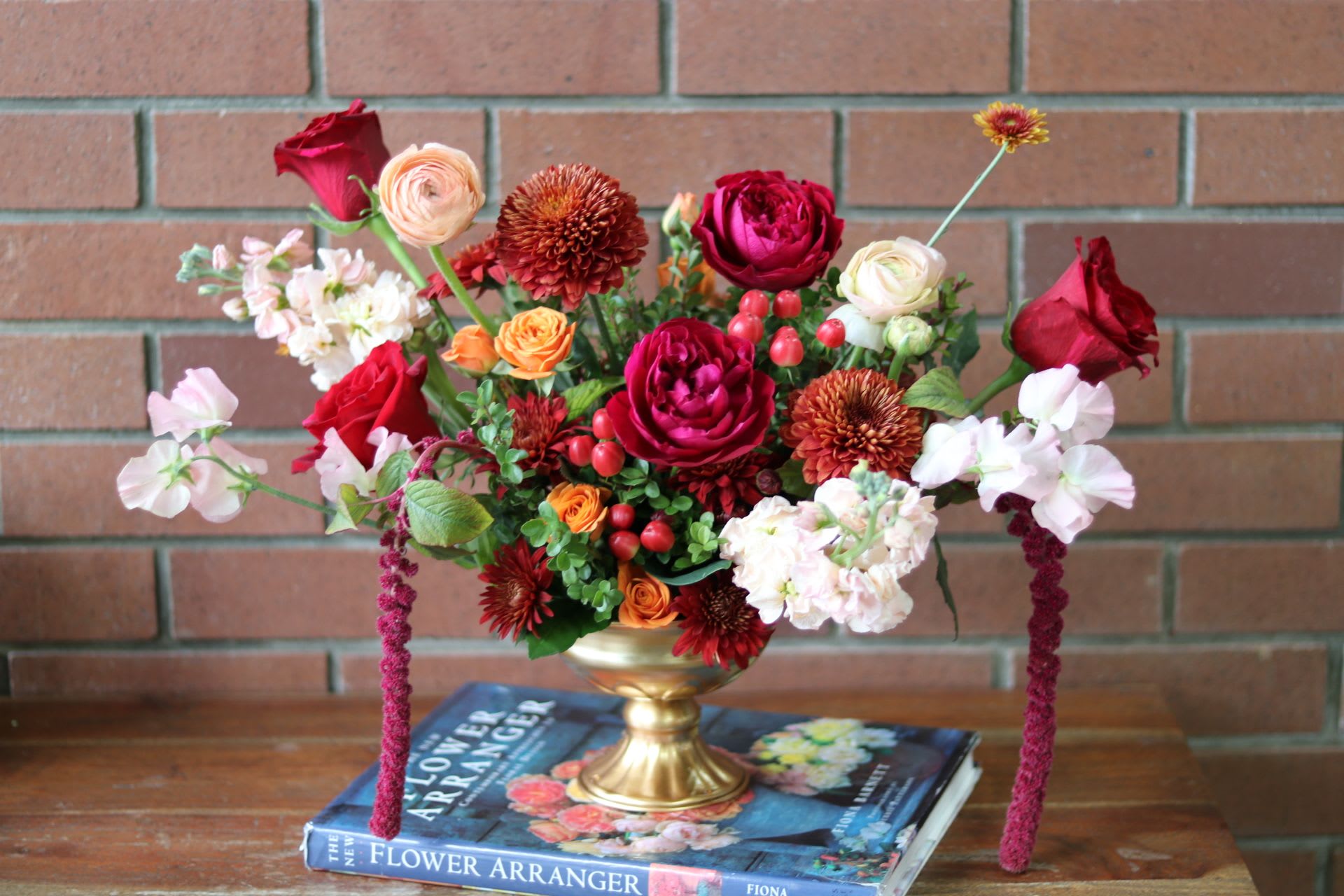 Golden Garden Elegance Centerpiece   - &quot;Golden Garden Elegance&quot;  The &quot;Golden Garden Elegance&quot; centerpiece is a stunning display of artistry and nature, designed in a luxurious gold compote that radiates opulence. At its heart, a lush arrangement of exquisite garden roses in deep red hues creates a romantic foundation. These are beautifully complemented by vibrant ball mums, delicate sweet peas, and charming ranunculus, each adding texture and depth. Sprays of fragrant stock and dainty spray roses weave throughout, enhancing the overall charm. The arrangement is artfully accented with an array of verdant greenery, providing a fresh and lively contrast that brings the whole piece to life. Perfect for Thanksgiving, special events, or as a striking focal point in any home, this centerpiece embodies the romance and beauty of a blooming garden.