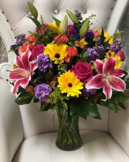 Celebrate Good Times Bouquet - This bouquet features a stunning mix of bold and cheerful blooms. It is anchored by large, star-shaped fuchsia and white stargazer lilies, complemented by a splash of bright yellow gerbera daisies. Deep magenta roses and clusters of purple stock flowers add rich texture and contrast. The arrangement is accented with orange spray roses and assorted greenery, all presented in a clear glass vase.