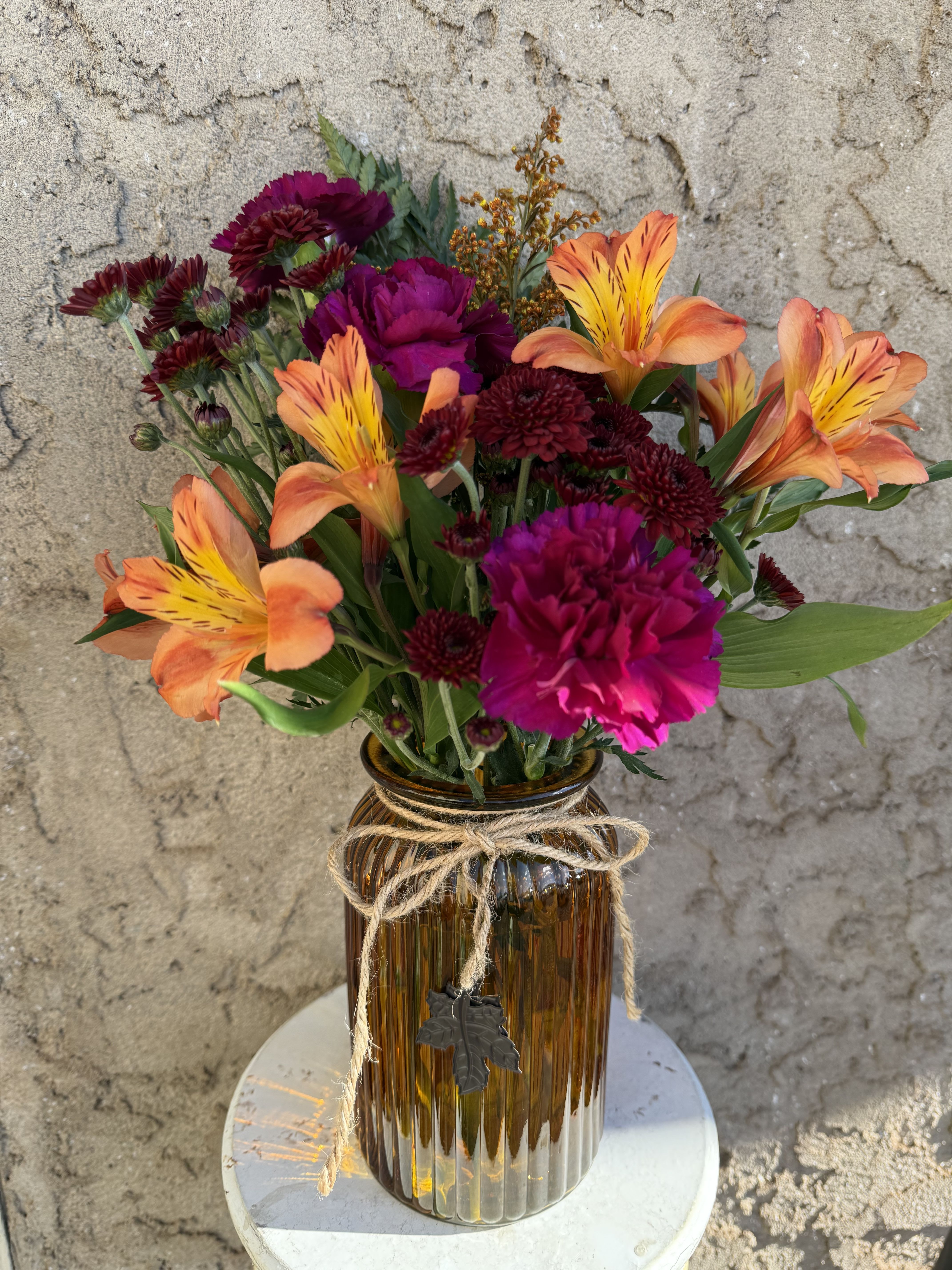 Copper Harvest - Vibrant and full of life, Copper Harvest combines bold autumn hues in a stylish ribbed vase tied with a gleaming metal leaf ribbon. Warm tones of burgundy, orange, and purple dance together in a cheerful medley of button mums, alstroemeria, carnations, and solidago. A radiant choice for fall decorating or gifting.  All flowers, floral colors, and containers are subject to change due to availability.
