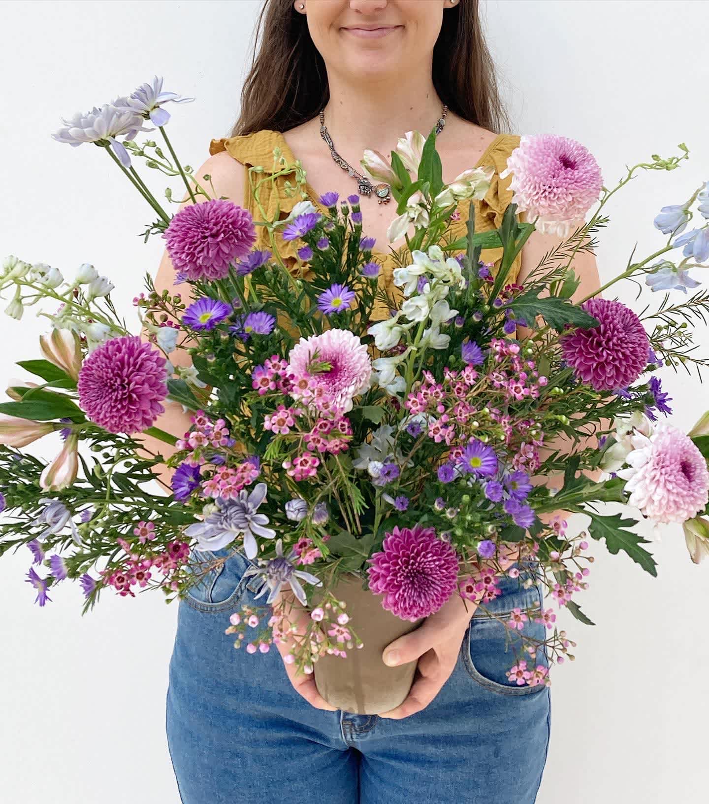 The Maeve - The Maeve is a serene and elegant floral arrangement, captivating with its soft hues and graceful presence. Pink wax flowers delicately weave through the gentle petals of purple mums and aster, creating a harmonious blend of color and texture. Airy light blue delphinium adds a touch of sophistication, beautifully complemented by light blue mums and pristine white alstroemerias. Perfect for any occasion, the Maeve radiates tranquility and refinement, making it a timeless choice for expressing heartfelt sentiments.