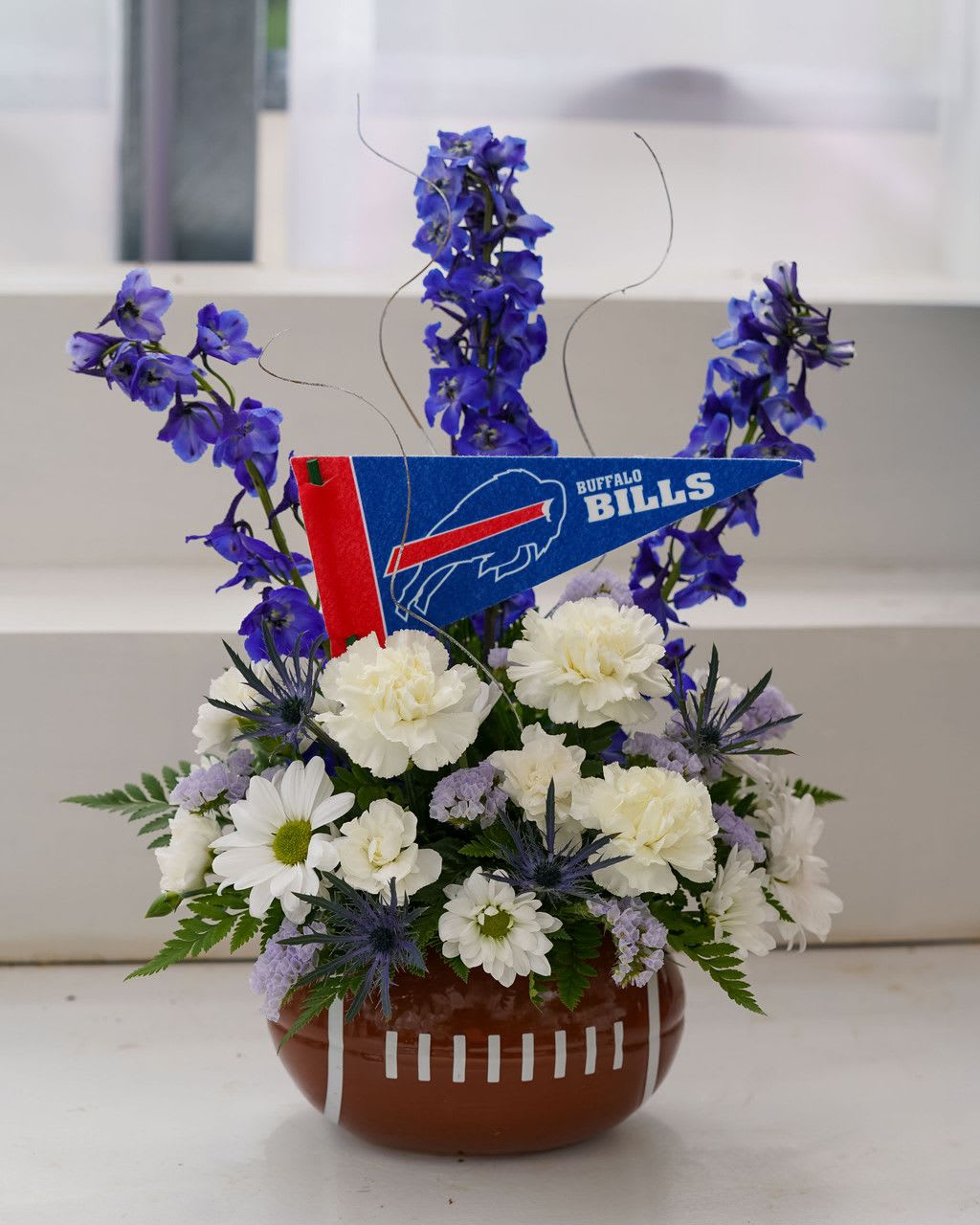 Cold Front Football Bouquet - Cold Front Football Bouquet  Inspired by the Buffalo Bills’ new Cold Front jerseys, this frosty arrangement brings the team’s bold spirit to life. Designed in a football keepsake container, it features icy white carnations, cool blue delphinium, crisp daisies, and frosty thistle, accented with a Bills pennant. Perfect for celebrating game day, gifting a die-hard fan, or showing off your team pride in style, just like the new look on the field.  PLEASE NOTE: Some flowers, vases and other products pictured are subject to availability, and may be substituted for a similar item if unavailable according to our substitution policy.