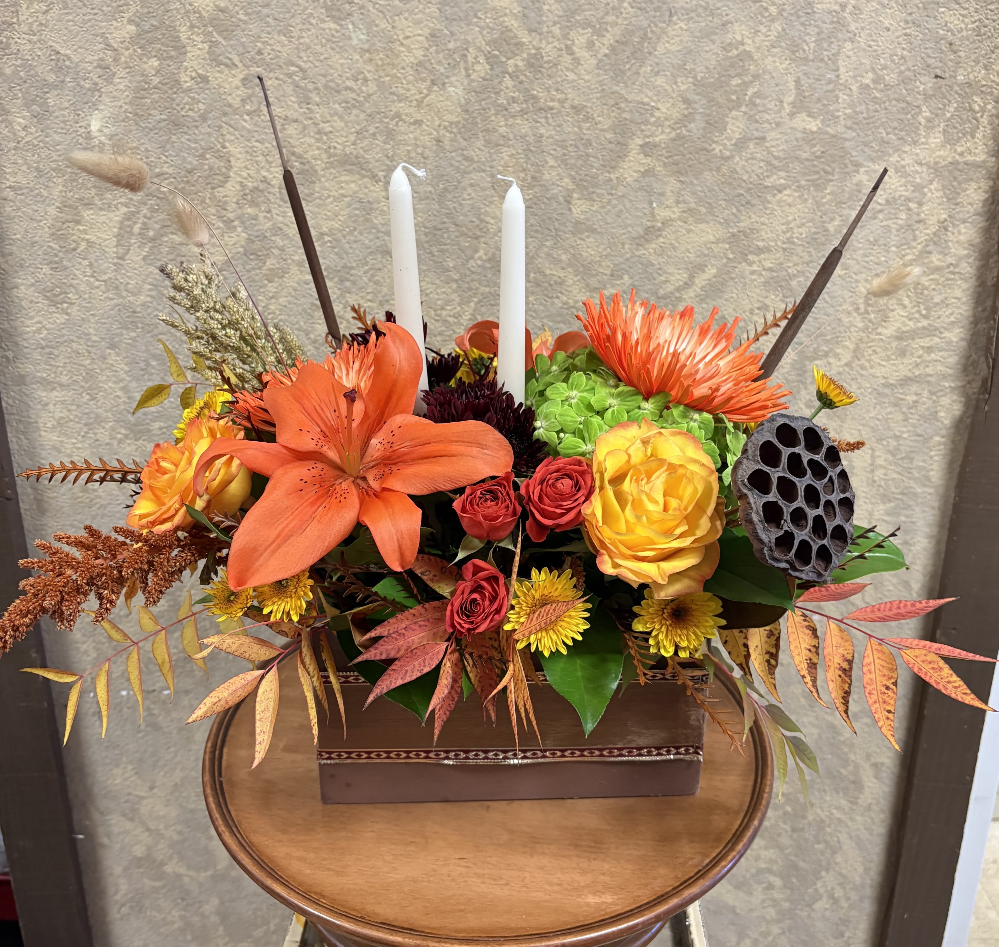 Gratitude and Thanks Centerpiece - Type of Flowers: Orange Roses, Orange Lilies, Orange Spray Roses, Fuji Mums, Daisies, and more with candle sticks in a brown wooden box planter. Availability: Autumn Design View: All Facing View Photo shown: Regular