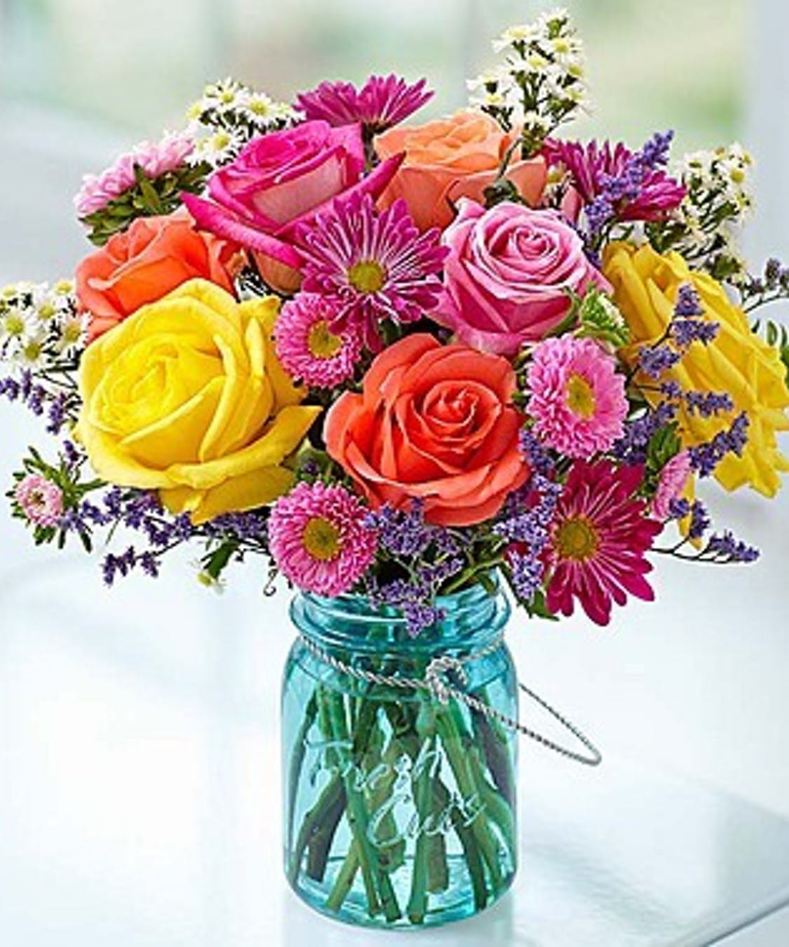 The Garden Jar - Capture the charm and color of the garden with this beautiful arrangement. Featuring vibrant roses, daisy poms, asters and more, and hand-designed in a reusable mason jar, it’s a gift that delivers endless smiles to all the special people in your life!  Arrangement of yellow, pink and orange roses, pink aster, daisy poms, purple limonium and white filler.