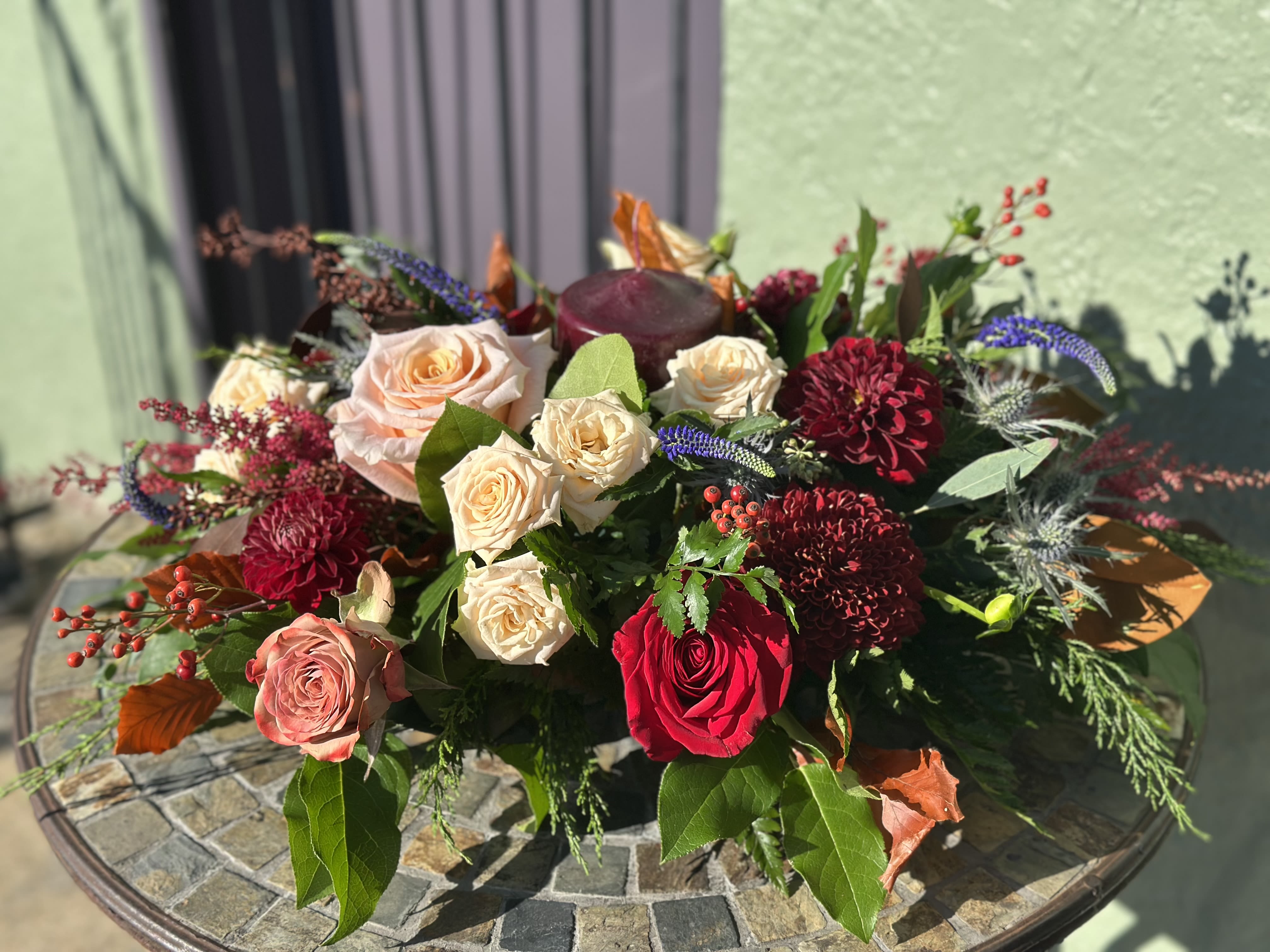 Fireside Gathering - Amber Gathering brings together the season’s richest hues in a lush, low centerpiece. Burgundy dahlias, cream, peach, and red roses mingle with soft blue veronica and textured seasonal foliage, creating a warm, inviting palette. Designed with a candle nestled at its heart, this centerpiece adds instant charm and cozy glow to dining tables and holiday gatherings.