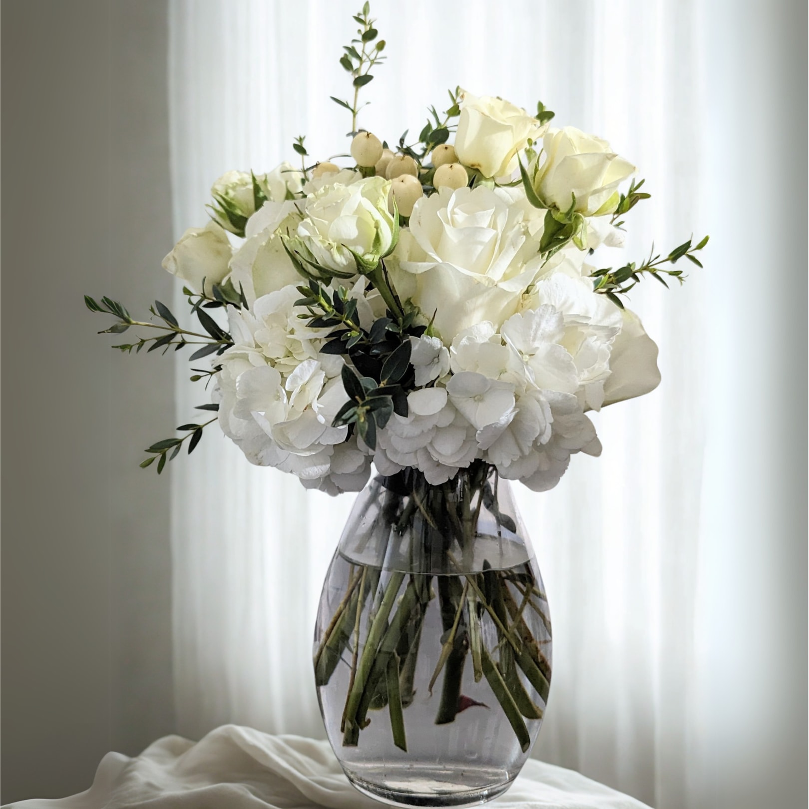 Pure Grace Vase - An elegant display of timeless beauty. This all-white arrangement features lush hydrangeas, premium roses, delicate lisianthus, and soft seasonal accents, gathered in a clear glass vase. Designed to evoke serenity and sophistication, it’s the perfect choice for sympathy, weddings, or as a refined gift for any occasion.