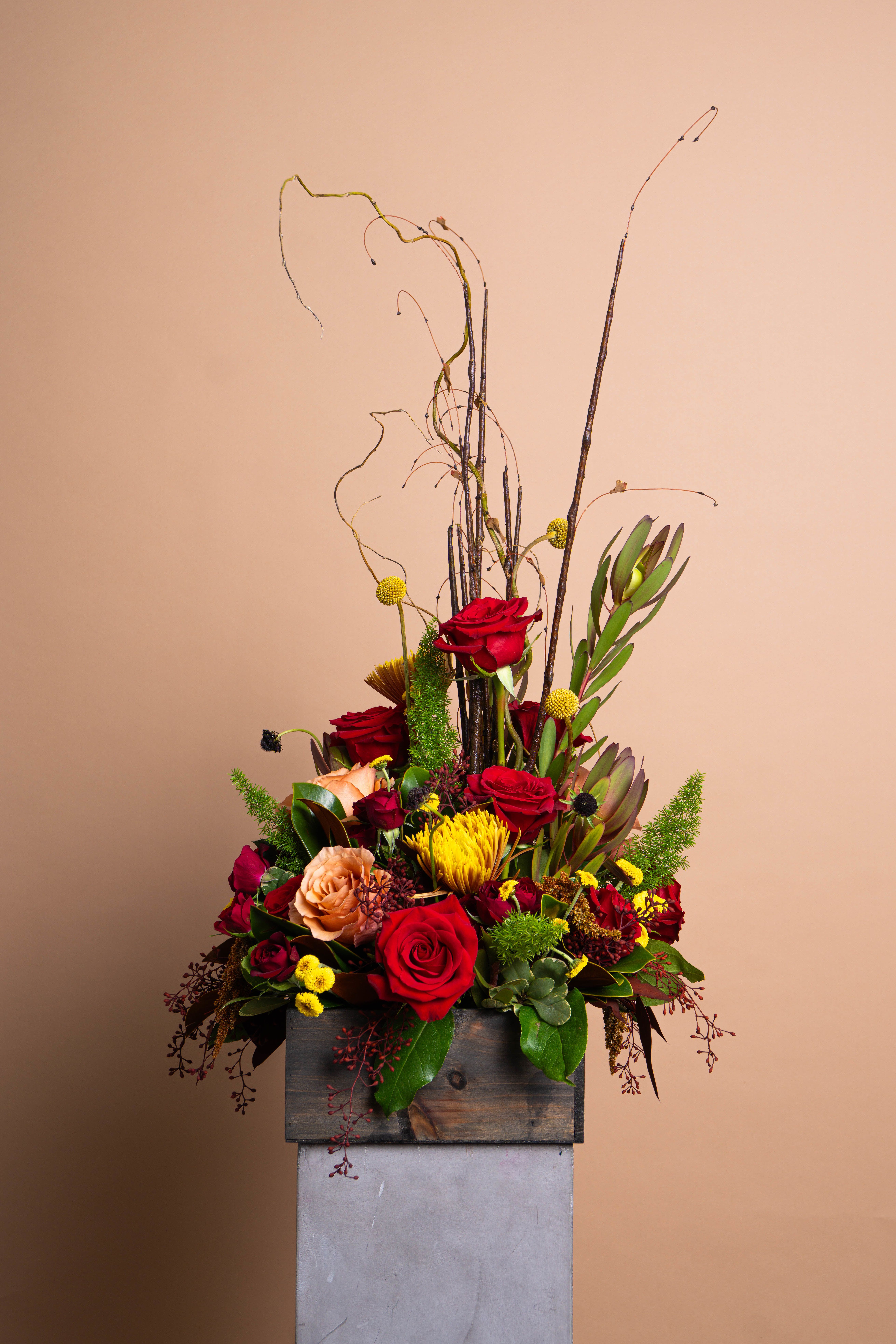 EMBER - Depth and heat in perfect balance. EMBER gathers crimson roses, toffee and golden blooms, and sculptural branches into a composition that feels both grounded and luminous. Natural textures meet modern structure, creating a piece that radiates quiet power.  Rich, layered, and elemental, EMBER captures the beauty of warmth held within stillness.  The arrangement pictured is Deluxe.