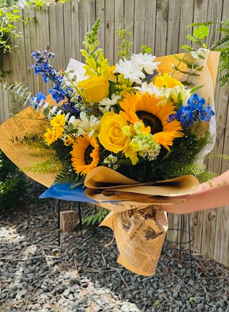 Ramblin' Road Wrap - Bursting with golden sunshine and blue-sky charm, this summer wrap captures the soul of a warm afternoon in a flower filled meadow. Bright sunflowers and cheerful yellow roses take center stage, their bold hues glowing against soft white daisies and delicate blue delphiniums. Wrapped in vintage-style newspaper, this hand-tied arrangement is perfect for porch sittin', lemonade sippin', or sending a little sunshine someone's way.