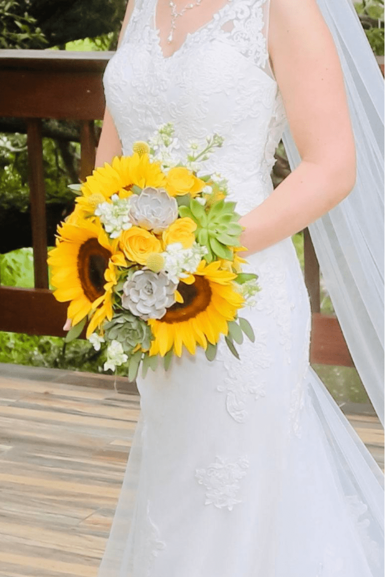 Jenna Sunflower Wedding Bouquet - Elevate your wedding with the Jenna Sunflower Wedding Bouquet. Stunning sunflowers, yellow roses, and succulents create a vibrant display, while white stock and fresh eucalyptus greens add a touch of elegance. Perfect for a whimsical, yet sophisticated affair.