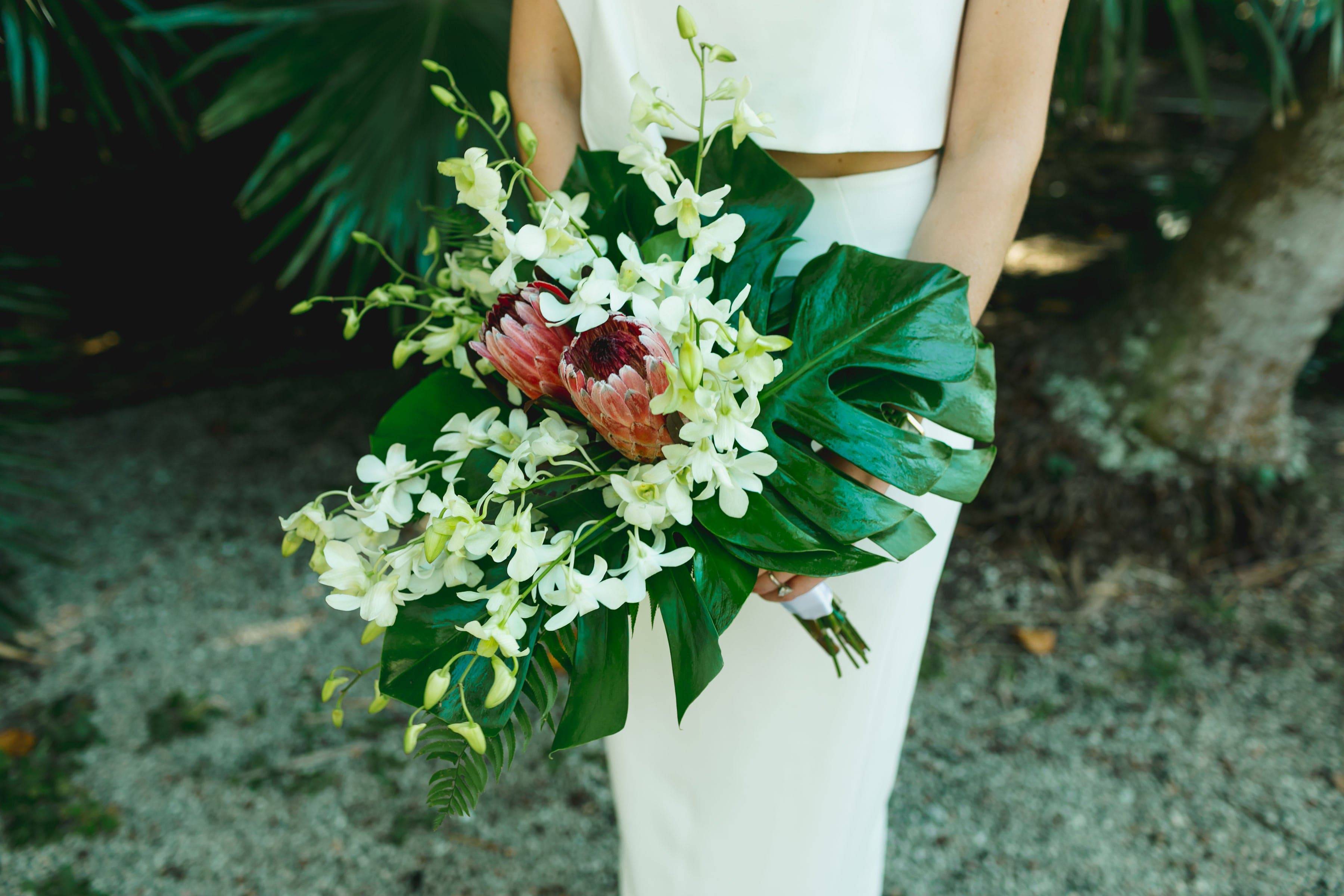 Whitney Tropical Wedding Bouquet - Indulge in the elegance of our Whitney Tropical Wedding Bouquet. Crafted with monstera leaves, white dendrobium orchids, and proteas, this exotic bouquet is the perfect accessory for a tropical themed wedding. Let these luxurious blooms transport you to paradise on your special day.