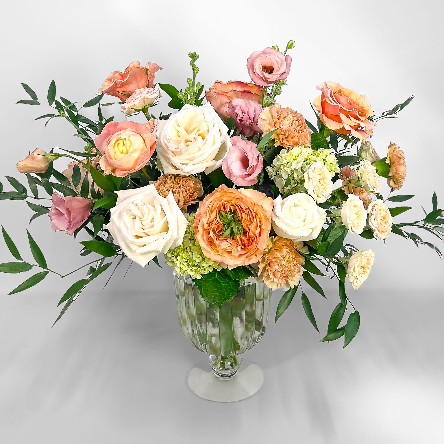Sunkissed - This garden-inspired arrangement features soft peach and pink blooms arranged with lush greenery. Presented in a charming footed vase, this arrangement exudes a welcoming, cozy vibe that radiates warmth and joy. Perfect for displaying in the home or sending as a thoughtful gift.