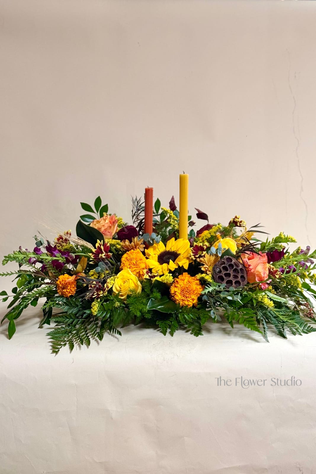 Autumn Frost  - Celebrate the warmth of Thanksgiving with this stunning Golden Harvest Centerpiece. Designed with rich fall blooms, including vibrant sunflowers, golden marigolds, roses, and accents of deep burgundy, it’s arranged with lush seasonal greenery and accented by autumn foliage and textured pods. Two taper candles in warm harvest tones complete the look, creating a cozy and inviting glow for your holiday table. Perfect for adding elegance and festive spirit to your Thanksgiving gathering.