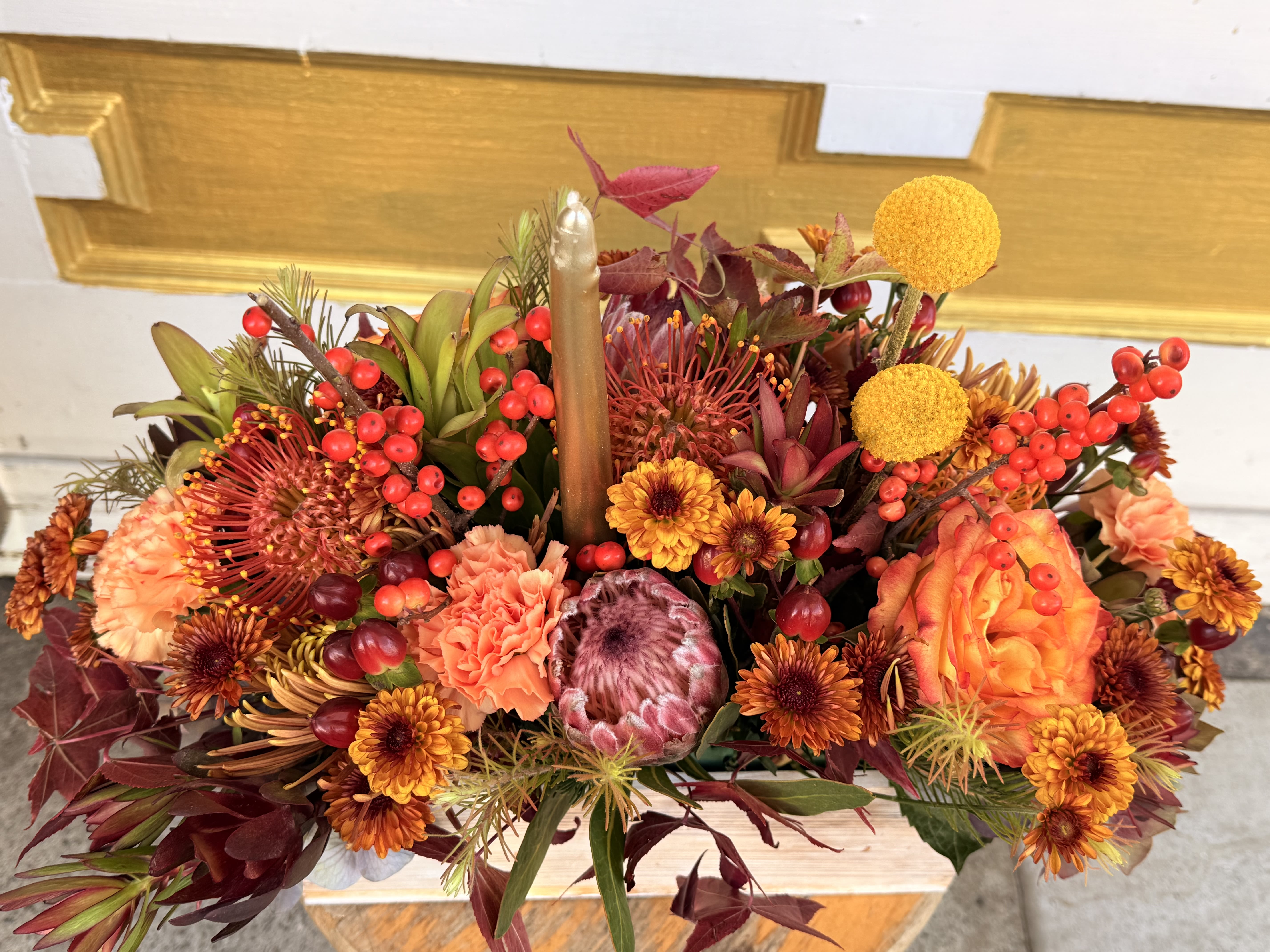 Golden Harvest ( table centerpiece ) - A luxurious autumn centerpiece bursting with golden warmth and texture. Featuring striking proteas, orange roses, bronze chrysanthemums, pincushion proteas, and golden craspedia, this design is complemented by clusters of berries and seasonal foliage in deep burgundy and amber hues. Nestled in a natural wooden box and finished with a gold taper candle, it radiates a cozy harvest glow — perfect for Thanksgiving tables or fall celebrations.