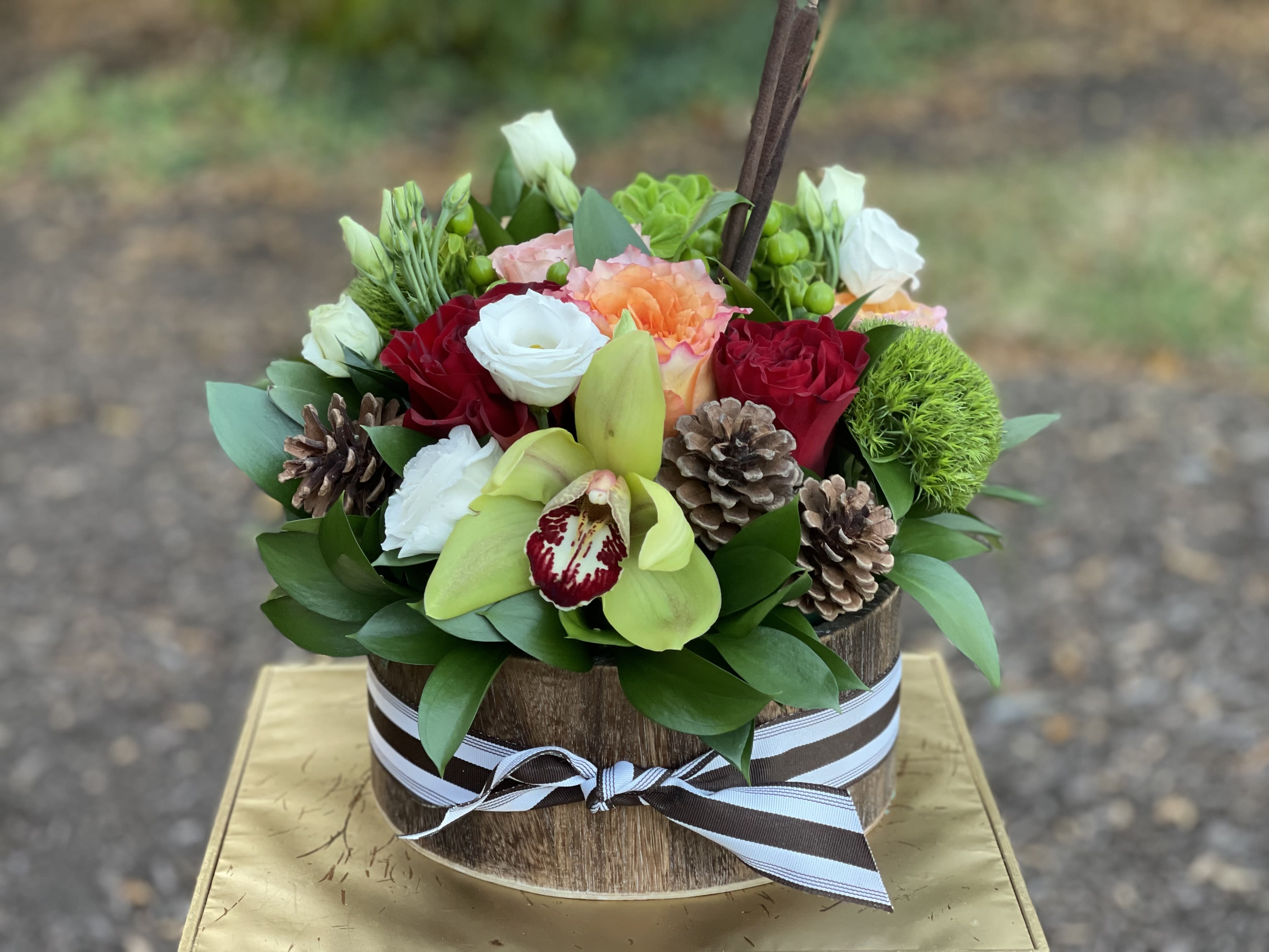 Pinecone Delight - Pinecone Delight | Wilmington, Delaware Florist Celebrate the cozy charm of the season with Pinecone Delight, a rustic yet elegant floral arrangement handcrafted by Petals Flowers and Fine Gifts in Wilmington, Delaware. This unique design features soft orange roses, rich red roses, delicate white lisianthus, lush green trichelium, exotic green orchids, and natural cattails, all accented with rustic pinecones for a warm, woodland-inspired touch. Arranged in a round wooden container, Pinecone Delight brings together seasonal textures and colors that feel both refined and inviting. Perfect for autumn celebrations, Thanksgiving tables, holiday gatherings, or as a heartfelt gift, this arrangement captures the beauty of nature with a sophisticated twist. Its earthy palette and natural elements make it an ideal centerpiece for homes, offices, or special events. As your trusted local florist in Wilmington, Delaware, we take pride in creating custom floral designs with the freshest blooms and premium accents. We offer same-day, tax-free flower delivery throughout Wilmington, Hockessin, Newark, Chadds Ford, Glen Mills, Kennett Square, and nearby communities, ensuring your flowers arrive fresh and full of beauty. Why Choose Petals Flowers and Fine Gifts? * Unique, luxury floral designs crafted with seasonal blooms * Family-owned Wilmington, Delaware florist with personal service * Same-day, tax-free flower delivery to local homes, offices, and event spaces