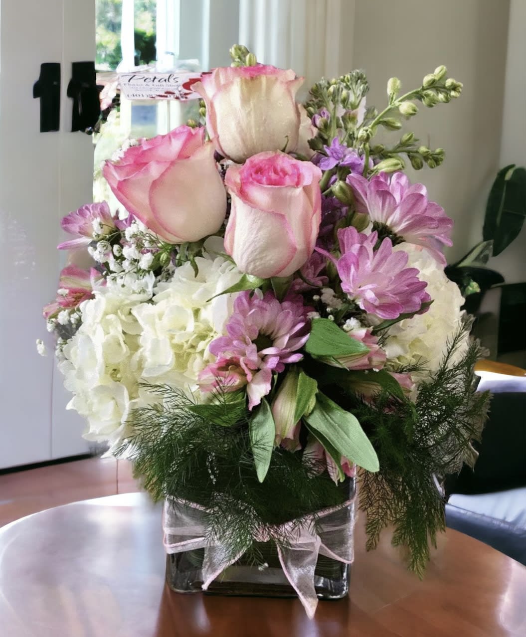Pinky Pink Cube by Petals -  This delightful floral arrangement is expertly designed within a stylish 5&quot; clear glass cube vase. It features a beautiful mix of pink roses, lavender chrysanthemum poms, Peruvian lilies, baby's breath, and lush greenery, creating a charming and &quot;pretty as can be&quot; display. It is sure to be a gift she loves! Please note: Rose colors may vary depending on daily availability and current chrysanthemum shades. Contact Us: For any questions regarding this specific design, please get in touch with Cheryl directly at 401-828-0701
