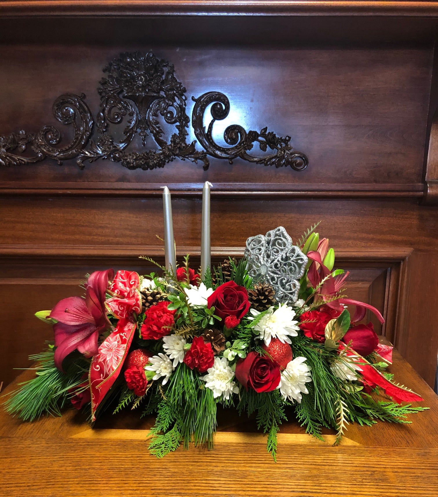 Christmas Centerpiece Red - Type of Flowers: Red Asiatic Lillies, Red Roses, Red Carnations, White Mums, and Pine greens, with two silver candles, pine cones, silver, and red ornaments and red ribbon in a plate. Availability: All year round Substitute Available: Yes Design View: All Facing View Photo shown: Regular