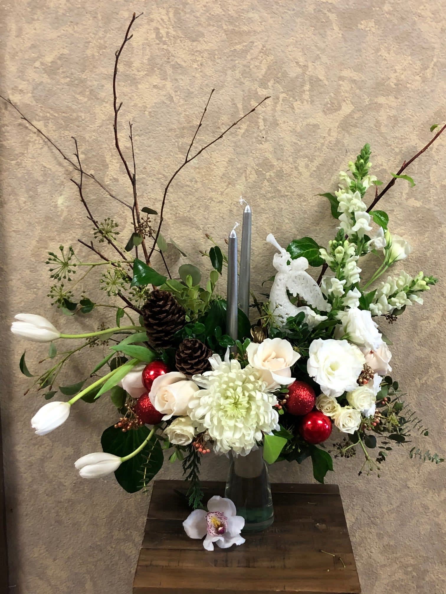 Angels Calling Christmas Centerpiece - Type of Flowers: White Snapdragons, White Dahlias, White Roses, White Tulips, White Lisianthus, Branches with two silver candles, pine cones, angel, red ornaments in a tall glass vase. Availability: All year round Substitute Available: Yes Design View: Front Facing View Photo shown: Regular