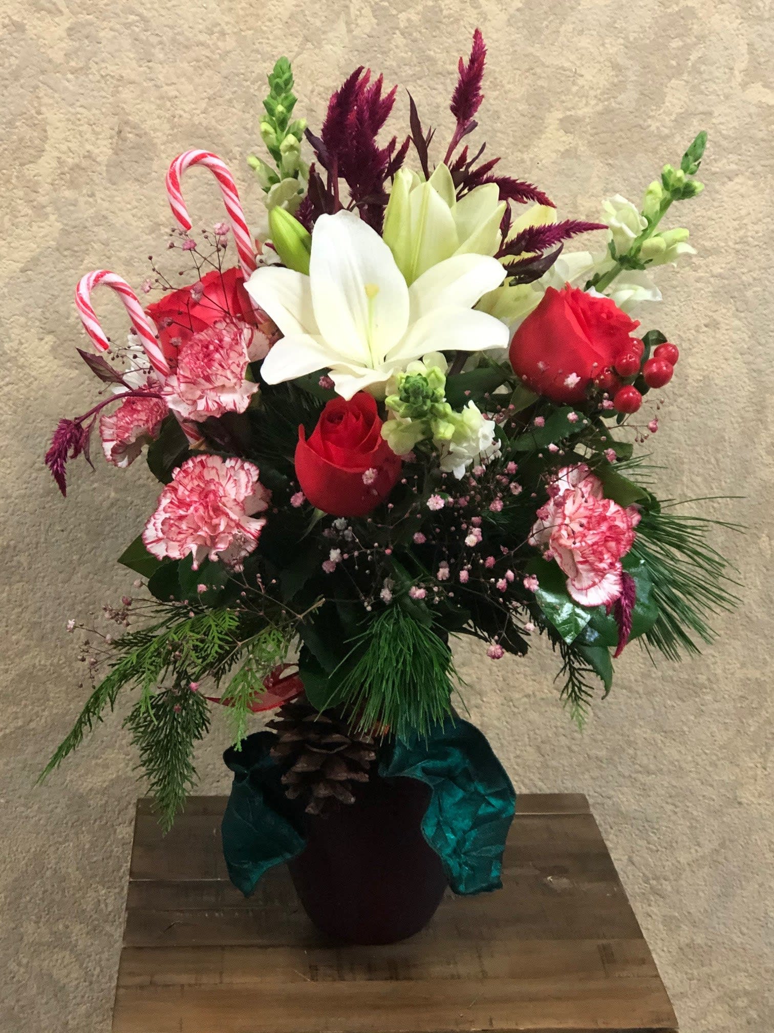 Holiday Cheer - Type of Flowers: White Snapdragons, Red Roses, White Asiatic Lilies,  Red Carnations, and pine greens in a red vase wrapped in teal ribbon and pine cones and moer. Availability: All year round Substitute Available: Yes Design View: Symmetric Front Facing View Photo shown: Regular