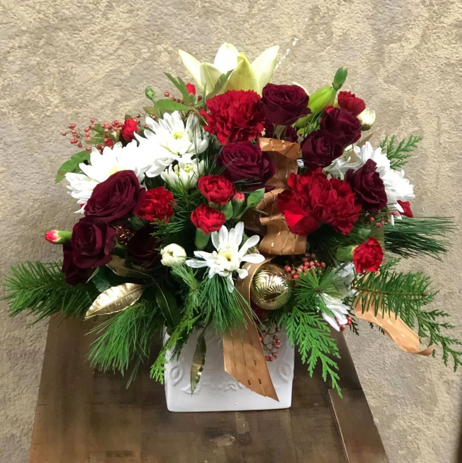 We Wish You a Merry Christmas - Christmas themed with red, whites, and golds. White asiatic lilies, red spray roses, white daisies, red mini carnations with a silver ribbon and ornaments in a white plaster. 