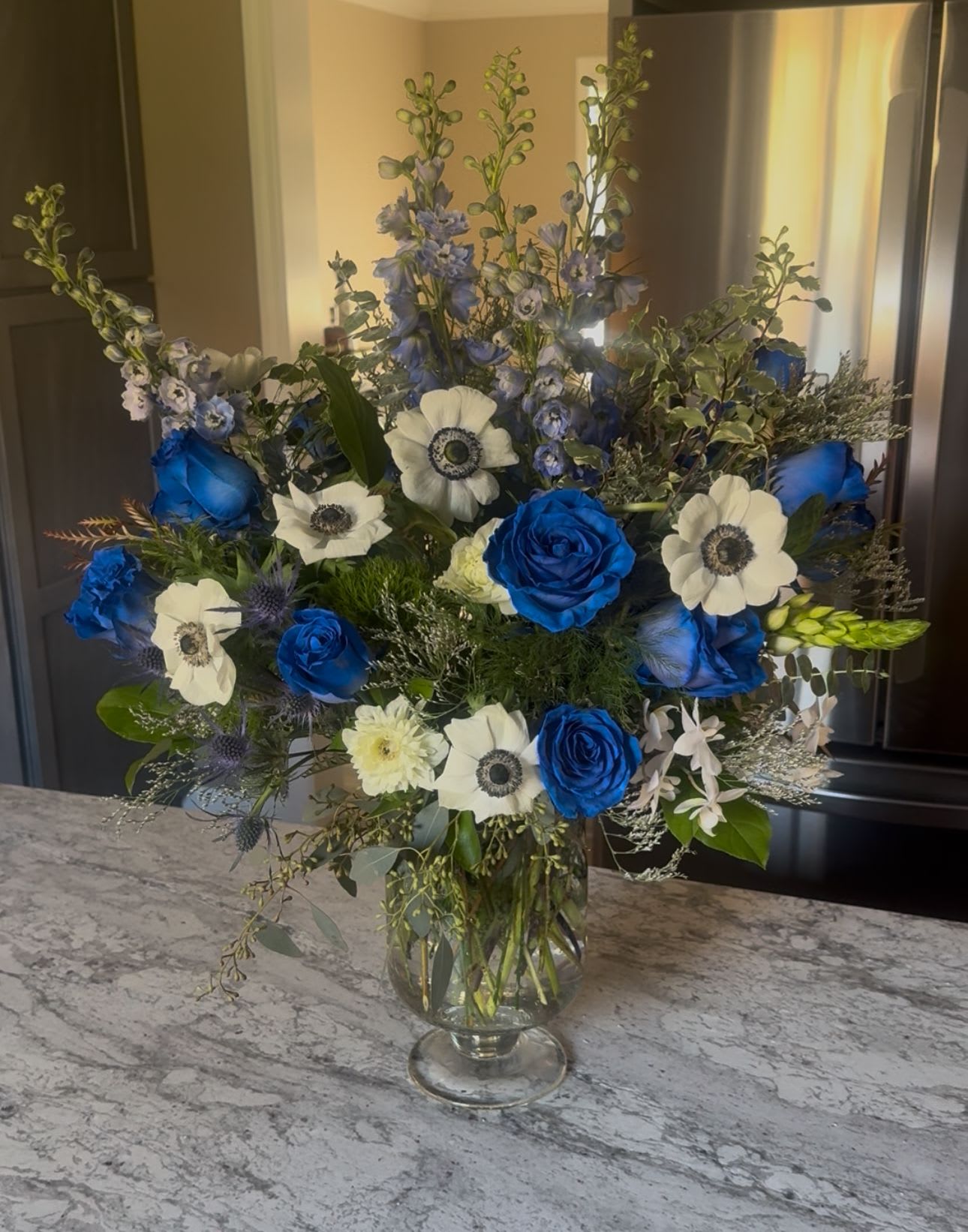 Blue Sway - Beautiful canvas of Woodland greenery, rich, Bellarosa blue roses, white Anemone, Israeli ruckus and Limonium. capturing blue suburban skies using baby blue Delphinium. This arrangement will capture true blue hearts!