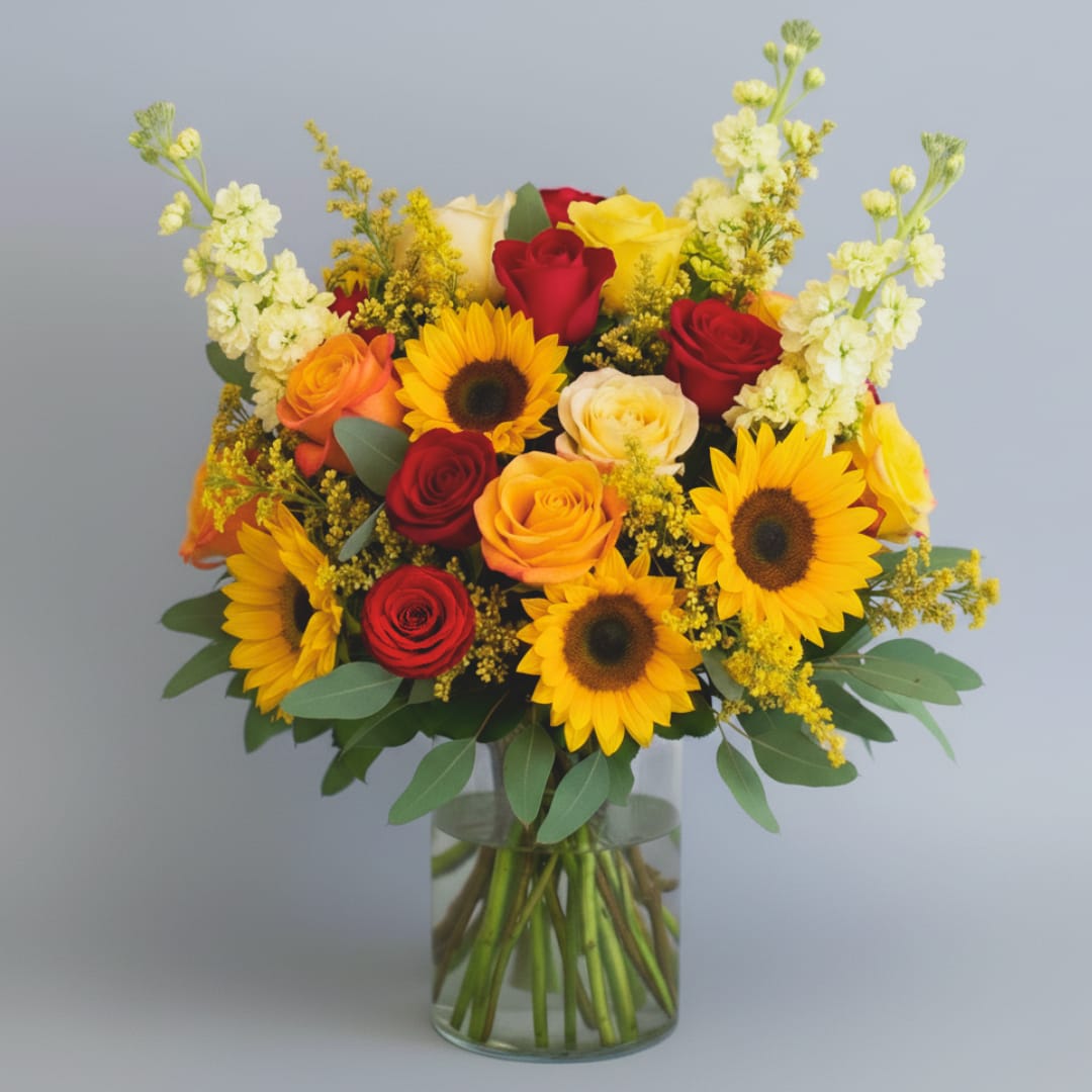 Autumn Romance - Autumn Romance  Embrace the cozy warmth and passionate hues of the season with our &quot;Autumn Romance&quot; bouquet. This richly textured arrangement is a lavish celebration of late summer and fall, perfectly capturing the feeling of a golden afternoon. Its full, rounded shape and varied textures create a look that is both deeply romantic and naturally abundant.  The heart of this design beats with the quintessential flowers of the season: bold, cheerful sunflowers instantly draw the eye with their dark centers and radiating yellow petals, symbolizing adoration and longevity. They are surrounded by an opulent mix of roses in colors that evoke sunset: deep crimson, bright red, and vibrant orange roses blend together to signify deep passion and heartfelt desire.  Adding height and a lovely, delicate contrast are creamy white spikes of stock flower, which lend a sweet fragrance and a graceful vertical element to the arrangement. Throughout the bouquet, delicate sprays of yellow solidago (or similar flower) weave in a soft, feathery texture and an extra layer of sun-drenched color.  Lush, blue-green eucalyptus foliage provides a relaxed, aromatic base, beautifully separating the warmer blooms while enhancing the overall garden-style feel. &quot;Autumn Romance&quot; is an expression of enduring beauty and deep affection, making it the perfect choice to convey warmth, happiness, and timeless love.  Arranged in a Cinched or Cylinder Glass Vase