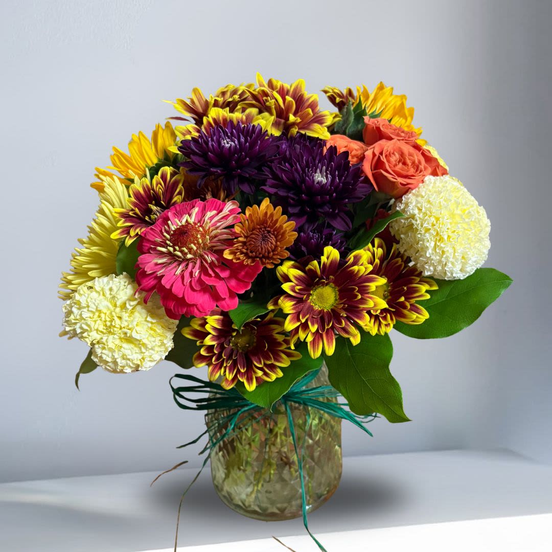 Friendsgiving Special - Type of Flowers: Seasonal Marigold, Seasonal Pink Zinnias, Fall Daisies, Orange Roses, Sunflowers, Chrysathemums, and Yellow Fuji Mums in an geometric clear vase with straw ribbon. Availability: Autumn Design View: All Facing View Photo shown: Standard