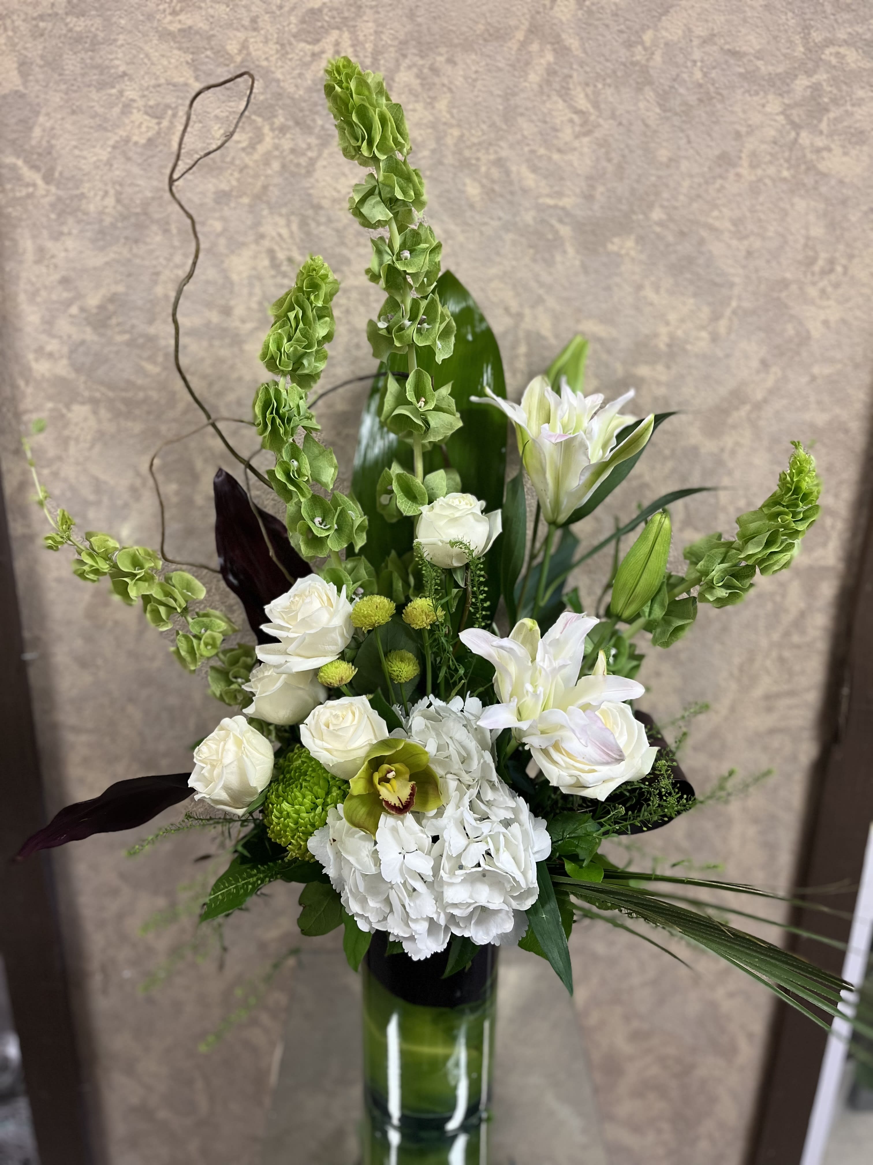 Emerald Isle, Ireland - Recipe: White Roses, White Asiatic Lily, Green Cymbidium Orchid, White Hydrangeas, Bells of Ireland, Green Chrysanthemums, Branches, and green leaves in a tall cylinder vase with a green leaf wrap. Availability: All year round Design View: Front Facing View Photo shown: Standard 