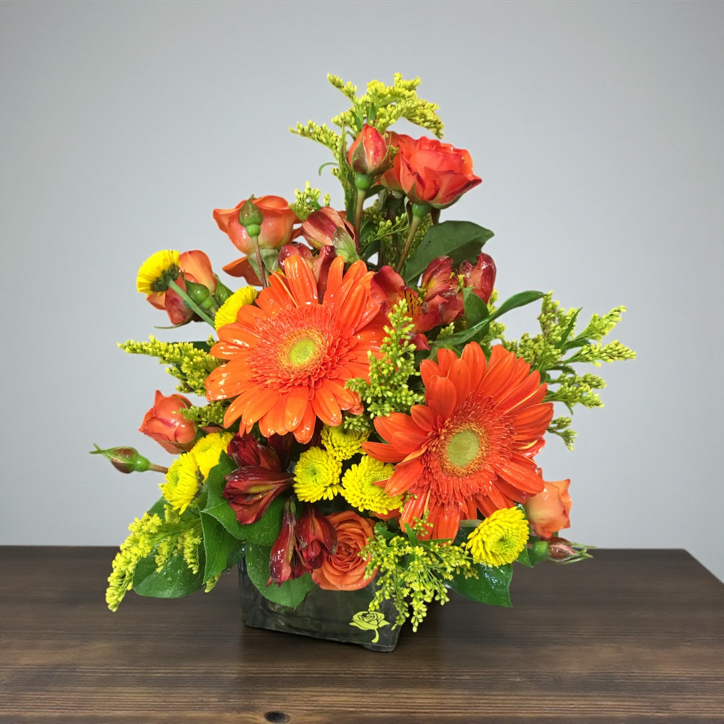 Grateful and Glowing - Bright, uplifting, and full of joy! This arrangement is the perfect way to send sunshine and gratitude in flower form. This cheerful arrangement features vibrant orange Gerber Daises, Rich spray roses, Sunny yellow buttons, and lushes greens, in a cube vase.