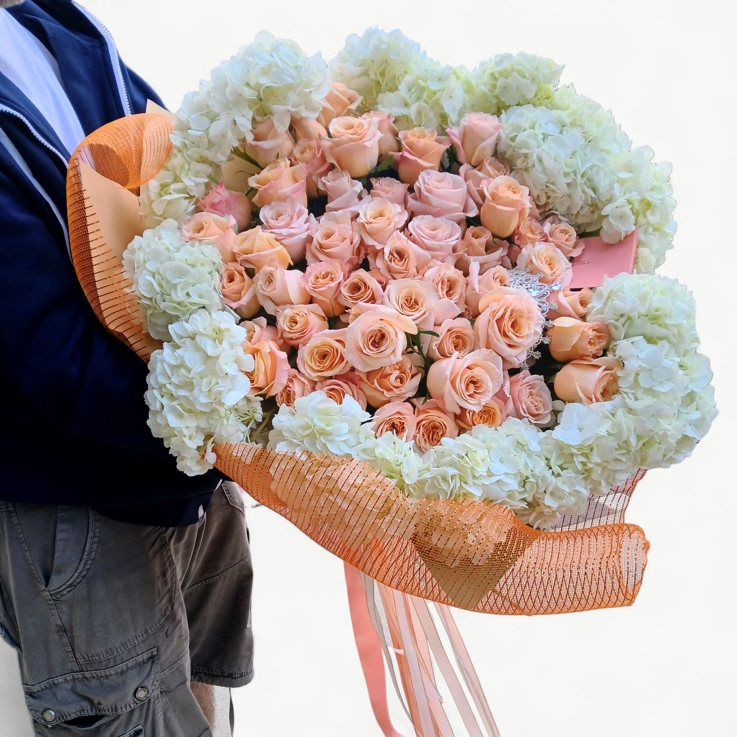 The Ultimate Luxury Grande Couture Rose Ring - Blush Peach Edition - A breathtaking statement of abundance and romance. This signature luxury Grande Couture Rose Ring is handcrafted with premium blush peach roses and surrounded by lush hydrangea for an unforgettable presentation.  Designed to make an entrance, this couture-scale bouquet is created completely by hand using the freshest luxury blooms of the day. Perfect for milestone celebrations, grand gestures, and moments that deserve the most extraordinary flowers. A real rhinestone couture tiara is included, and can be worn of displayed as a keepsake.  Every piece is designed exclusively by Joey-Lynn, using her signature Italian-inspired style, artistry, and attention to exquisite detail. Please allow 48 hours advance notice for this couture design so we may source the most exceptional blooms.