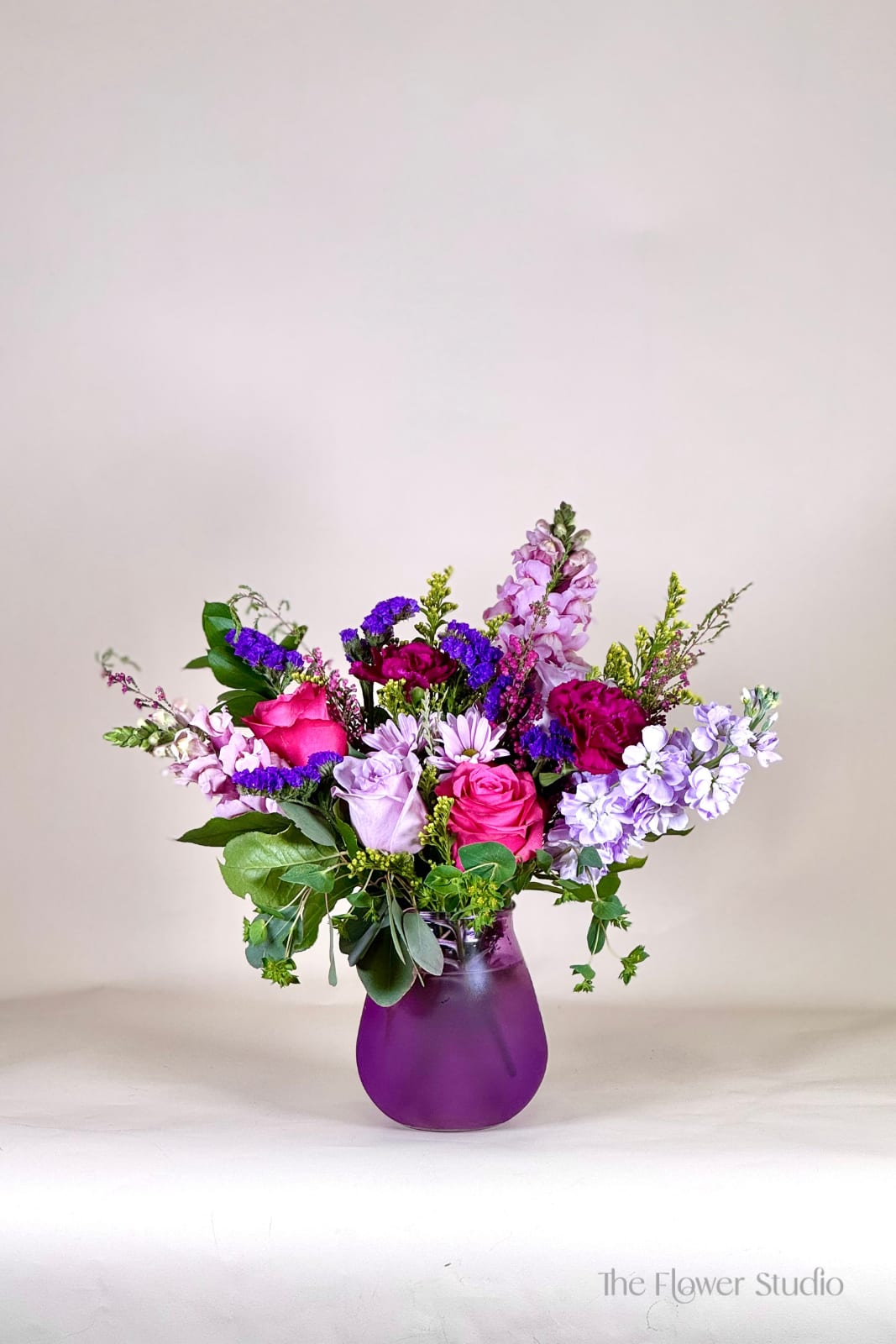 Purple Heart - A charming and elegant floral arrangement designed in rich purple hues, perfect for any occasion. This petite yet stunning bouquet features a blend of lush seasonal blooms, such as delicate lavender and pruple blooms, all beautifully arranged in a stylish purple glass vase. Accented with wisps of greenery for a natural touch, this low and compact design is ideal for tabletops, desks, or as a heartfelt gift. Whether celebrating a special moment or simply brightening someone's day, Purple Heart brings a sense of calm, beauty, and timeless elegance to any space.  Our floral artists hand-design each arrangement, so flower and container styles may vary based on local and seasonal availability. As a result, we are unable to guarantee specific florals, colors, or vessels—but we always ensure the same level of beauty and value in every piece.